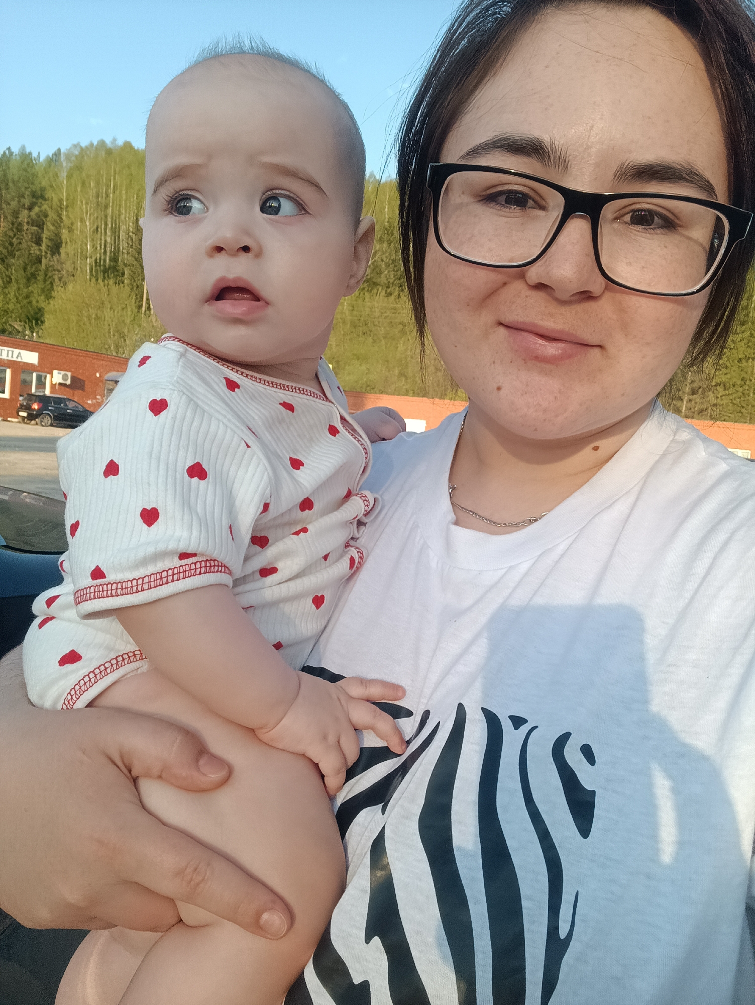A woman wearing glasses and a white t-shirt with a zebra print is holding a baby. The baby is wearing a white onesie with red hearts. In the background is a building with the word "ТПА" visible, and a vehicle parked nearby. Trees are visible in the background. The location is likely Uinskoye, Russia, based on the Cyrillic inscription on the building.