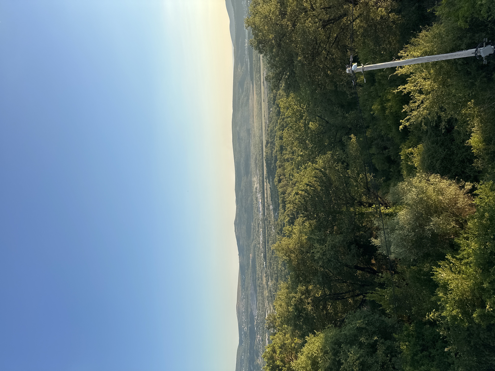 This is a vertical, eye-level, outdoor photograph taken from a high vantage point overlooking a valley and distant hills under a clear blue sky. In the foreground, lush green trees and vegetation dominate the lower portion of the frame. A utility pole with electrical equipment is visible on the right side of the image, emerging from the dense foliage.
The mid-ground features a valley with scattered buildings, roads, and what appear to be industrial or quarry sites. The valley is surrounded by rolling, tree-covered hills that recede into the distance. The horizon is marked by a range of mountains, softened by atmospheric haze. The light suggests it is late afternoon or early morning, with the sun casting a warm glow on the landscape.
The scene is part of the natural landscape and infrastructure surrounding the city of Kavač, Montenegro, characterized by its hilly terrain and proximity to the coast. There are no people or animals visible in the photograph.