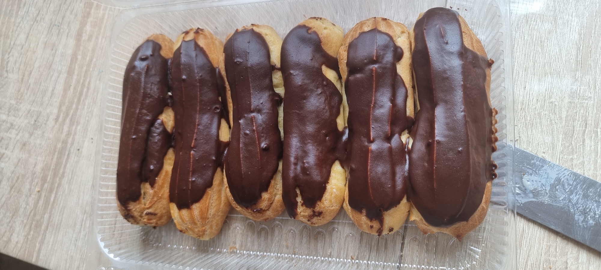 A clear plastic container holds six chocolate-glazed eclairs arranged in two columns of three. The eclairs are positioned on a light-colored wooden surface, which appears to be a table. The lighting is bright, casting soft shadows. A portion of a metal knife with a white residue is visible in the lower left corner, suggesting it may have been used to cut or prepare the eclairs. The context of Aktobe, Kazakhstan is not discernible from the visual information in the image.