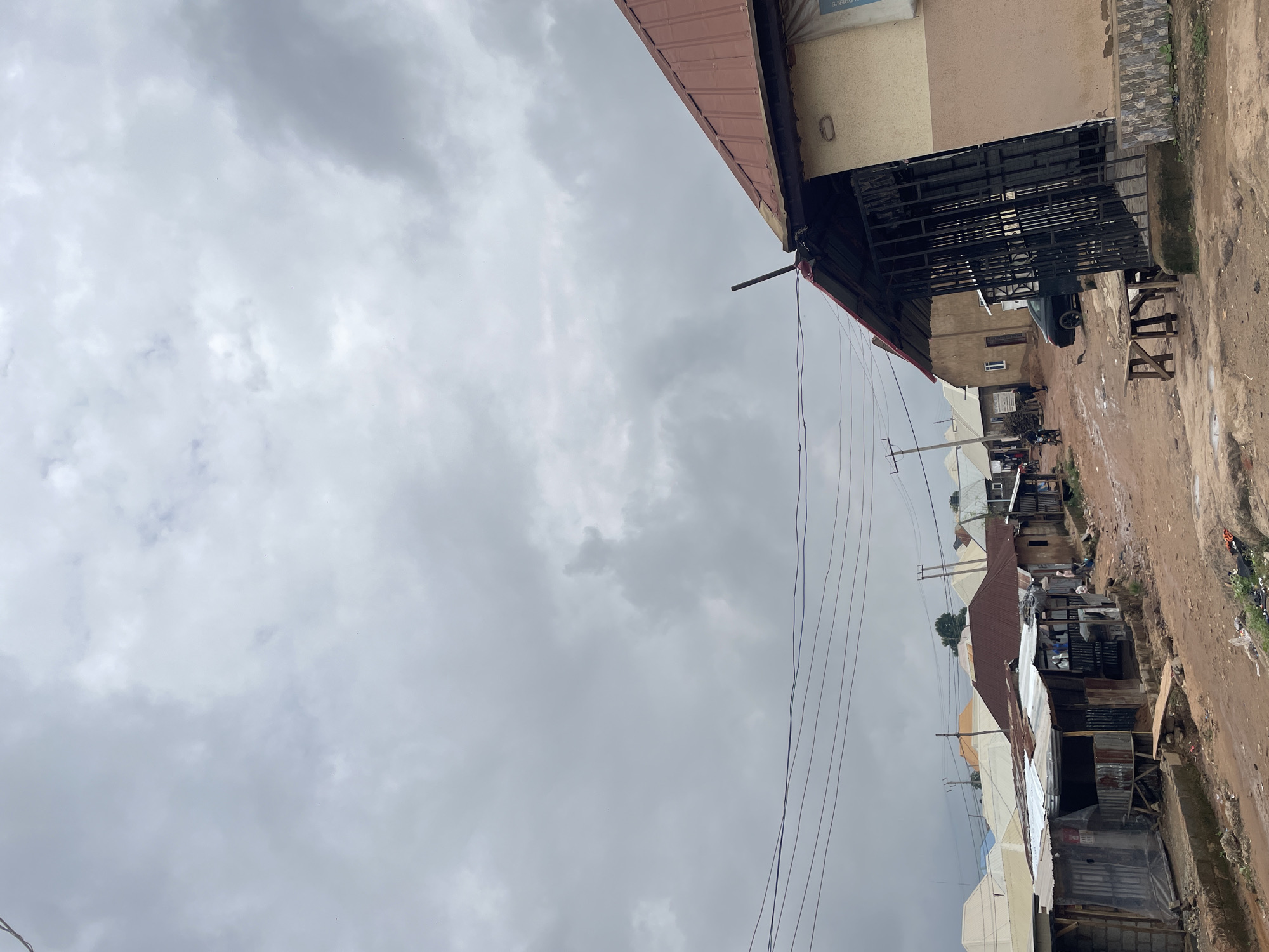 This image depicts a street scene in Kurunduma, Nigeria. The sky is overcast with dense grey clouds. In the foreground, a dirt road is wet and muddy, indicating recent rain. On the right side of the road, a building with a red corrugated metal roof and light-colored walls is visible, featuring a black metal gate. A small wooden table and bench are positioned in front of the building.
Further down the street, several other buildings with corrugated metal roofs in various states of repair are visible. Some are constructed with corrugated metal sheeting and what appear to be cinder blocks. In the middle ground, a black car is parked on the road. A few individuals are present, including some walking and possibly interacting with one of the structures. Electrical wires are strung across the scene. The overall impression is of a modest, developing urban environment.