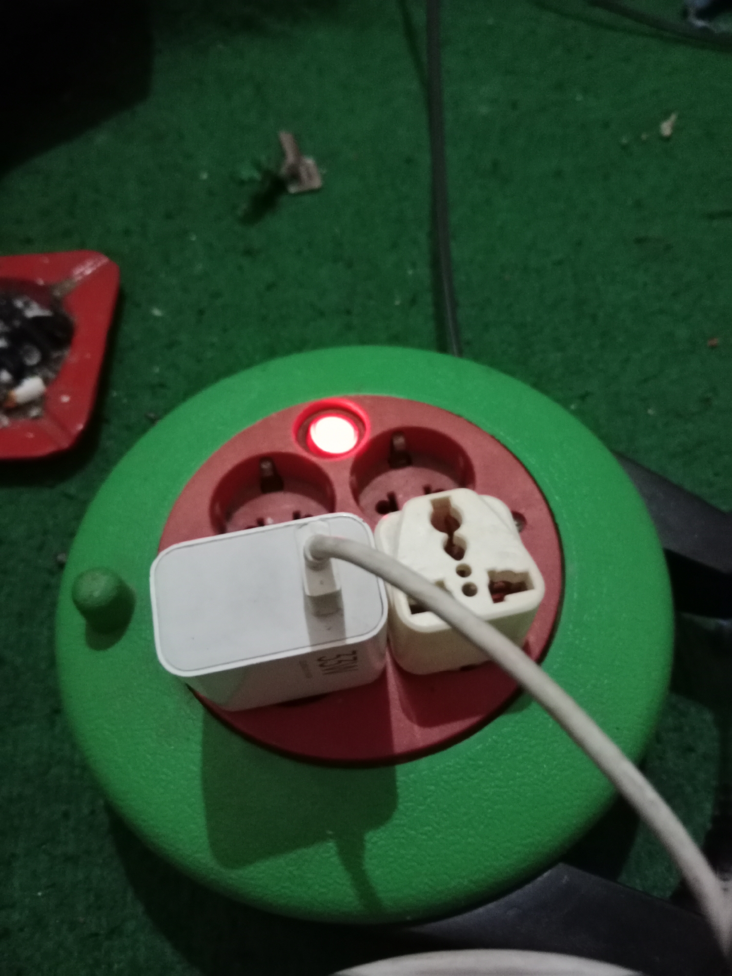 In Ringinrejo, Indonesia, a close-up shot focuses on a green and red power strip lying on a green carpeted surface. The power strip features three sockets, one of which is illuminated by a red indicator light, suggesting it is active. Two electrical adapters are plugged into the sockets. The left adapter is white and rectangular, likely a power brick, with a white cable emerging from it. The right adapter is a universal multi-socket adapter, also white, with multiple slots for different plug types. A thick gray cable extends from the power strip, disappearing out of frame. To the left of the power strip, a red ashtray containing cigarette butts and other small debris is visible. The lighting in the scene is dim, casting shadows on the carpet.