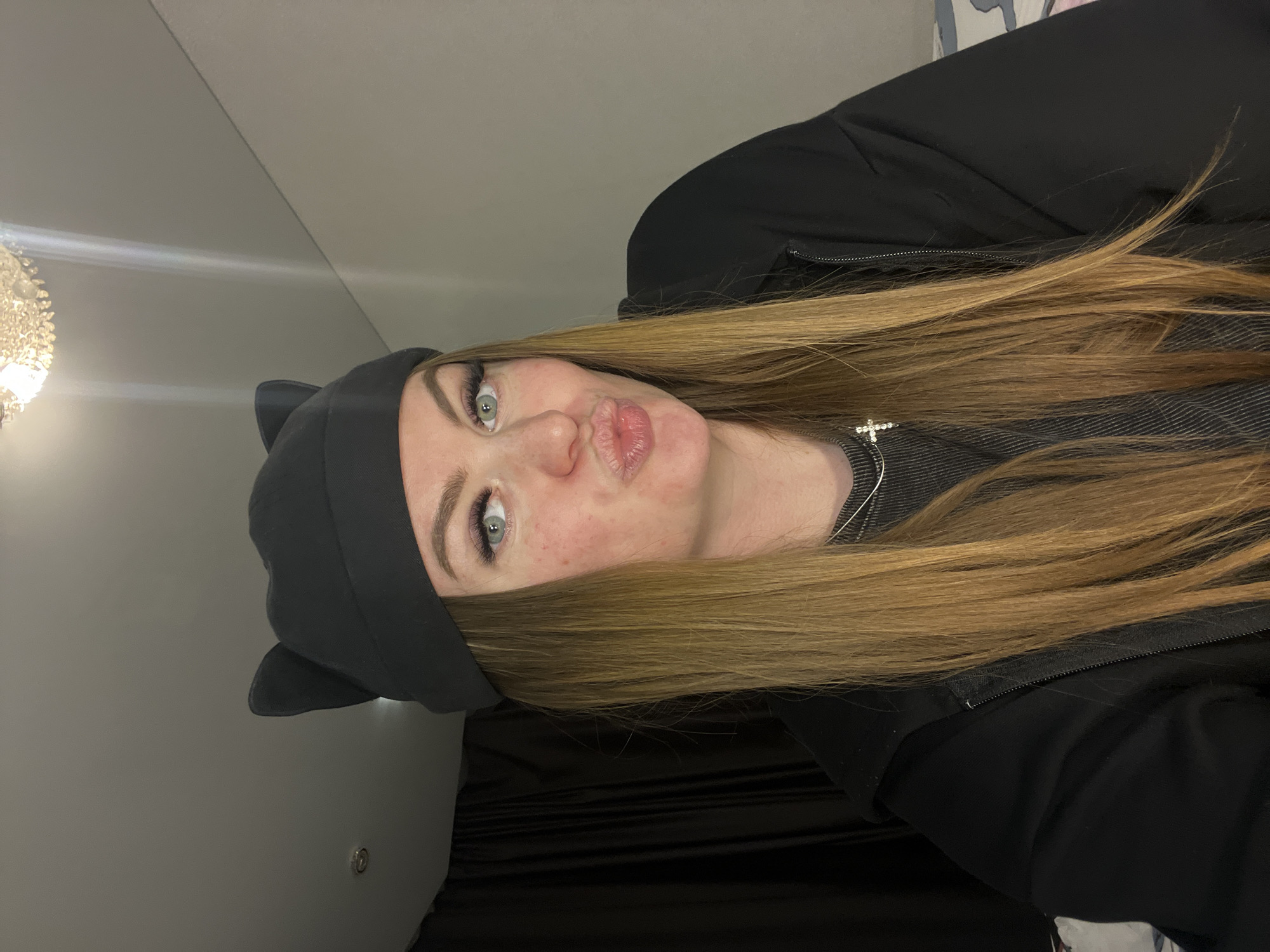 A selfie of a young woman, possibly in an indoor environment. She is wearing a black jacket and a gray beanie with cat ears. She has long blonde hair and is wearing a silver necklace with a cross pendant. She is making a pouting face. The background is partially visible, showing a wall with a light fixture and dark drapes. The location is Unknown, Unknown.