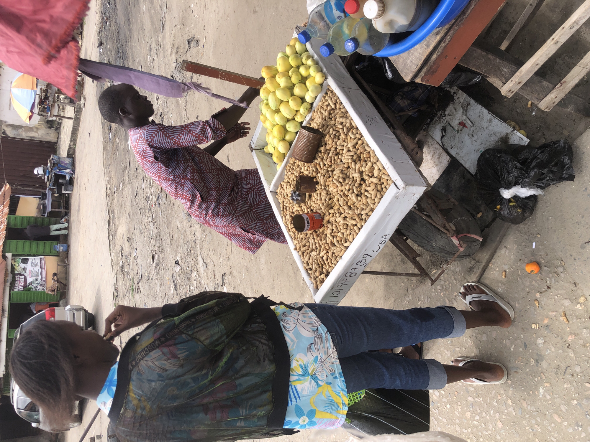 In Ado, Nigeria, a street vendor displays a cart overflowing with produce. The cart, a white wooden structure, is divided into two compartments, one filled with a large quantity of peanuts in their shells, and the other with bright yellow, elongated fruits, possibly mangoes or papayas. Two empty tin cans sit atop the peanuts, one with a faded red label.
A young woman, her back to the camera, wears a colorful patterned jacket over a floral print top and blue jeans. She is eating something from her hand, her fingers touching her mouth. To her right, a young man in a red and white patterned tunic leans over the cart, reaching towards the produce.
Behind the cart, a woman with a large red umbrella stands under a makeshift stall. Several bottles of drinks, some clear and some with blue or red caps, are visible near the front of the cart. The ground is dusty and unpaved, with scattered debris and a few small orange objects that could be discarded fruit. In the background, a silver minivan and a building with green signage that reads "MAMA G'S KITCHEN" suggest a bustling urban environment. The overall scene depicts a typical day at a Nigerian street market.