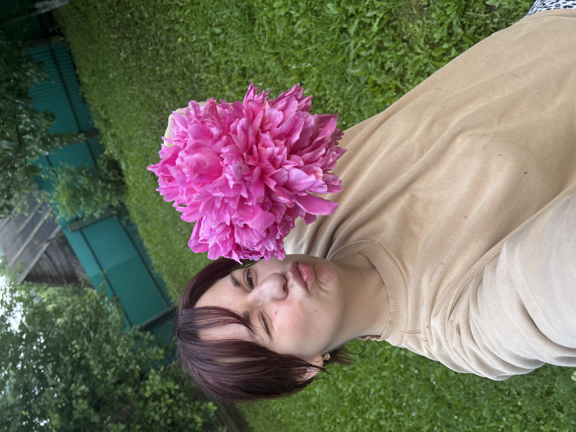 A woman with dark hair and bangs winks and pouts while holding a large, vibrant pink peony in front of her face. She is wearing a tan sweatshirt. The background is a lush green lawn with a teal fence and some trees, suggesting an outdoor setting in a rural area, potentially the city of Fokino, Russia.