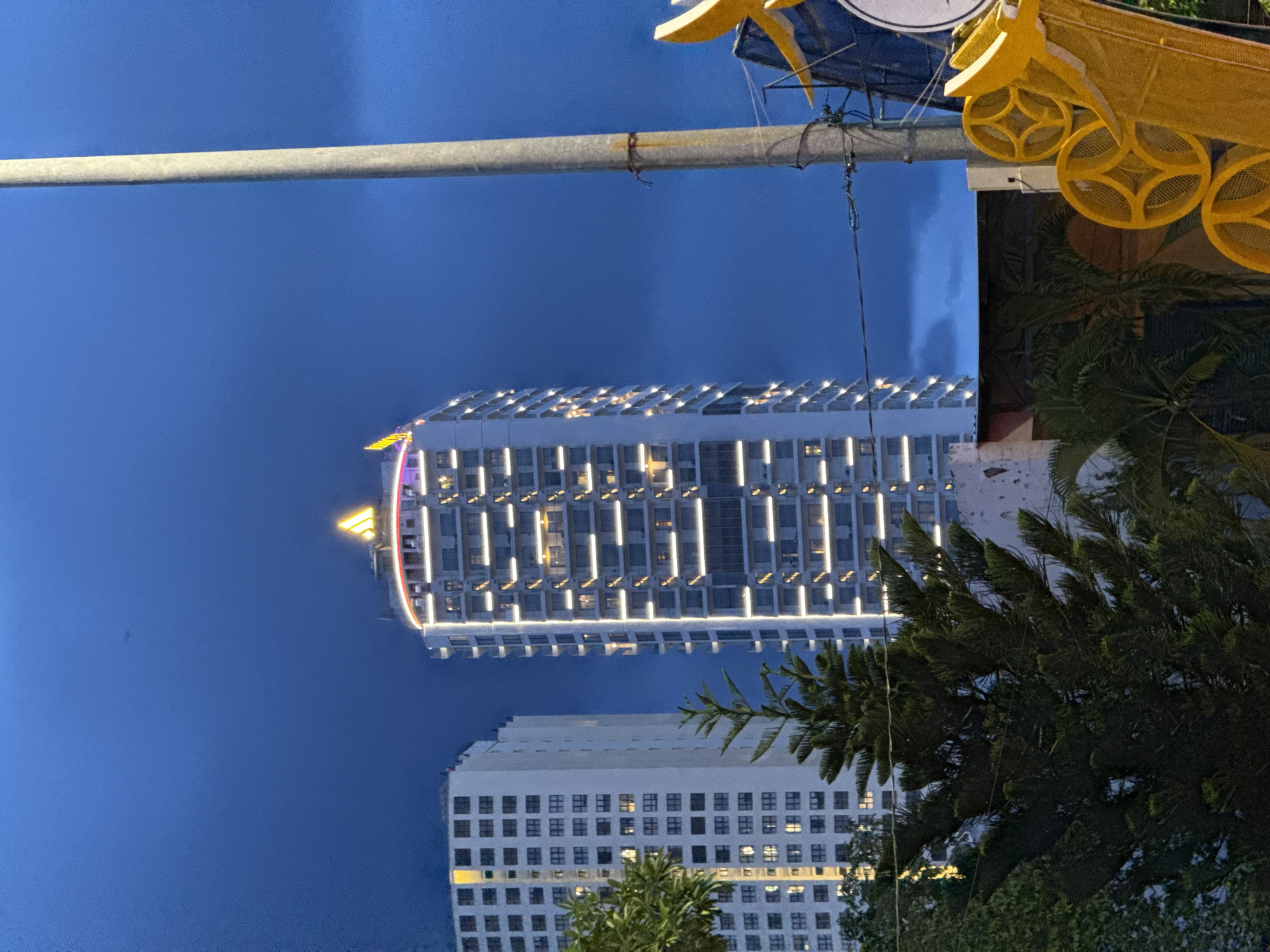 In the twilight of Nha Trang, Vietnam, two towering buildings dominate the scene against a deep blue sky. The building on the right, a modern skyscraper, is illuminated with a vibrant cascade of white lights tracing its vertical lines and balconies, culminating in a bright yellow, triangular logo at its peak. To its left stands a second, similarly tall building, also white, with rows of illuminated windows. In the foreground, a lush evergreen tree, likely a Norfolk Island pine, and the fronds of palm trees create a natural counterpoint to the urban architecture. A utilitarian light pole stands to the right of the frame, its gray concrete texture contrasting with the sleek lines of the buildings. The overall impression is one of urban development meeting natural elements under the fading light of day.