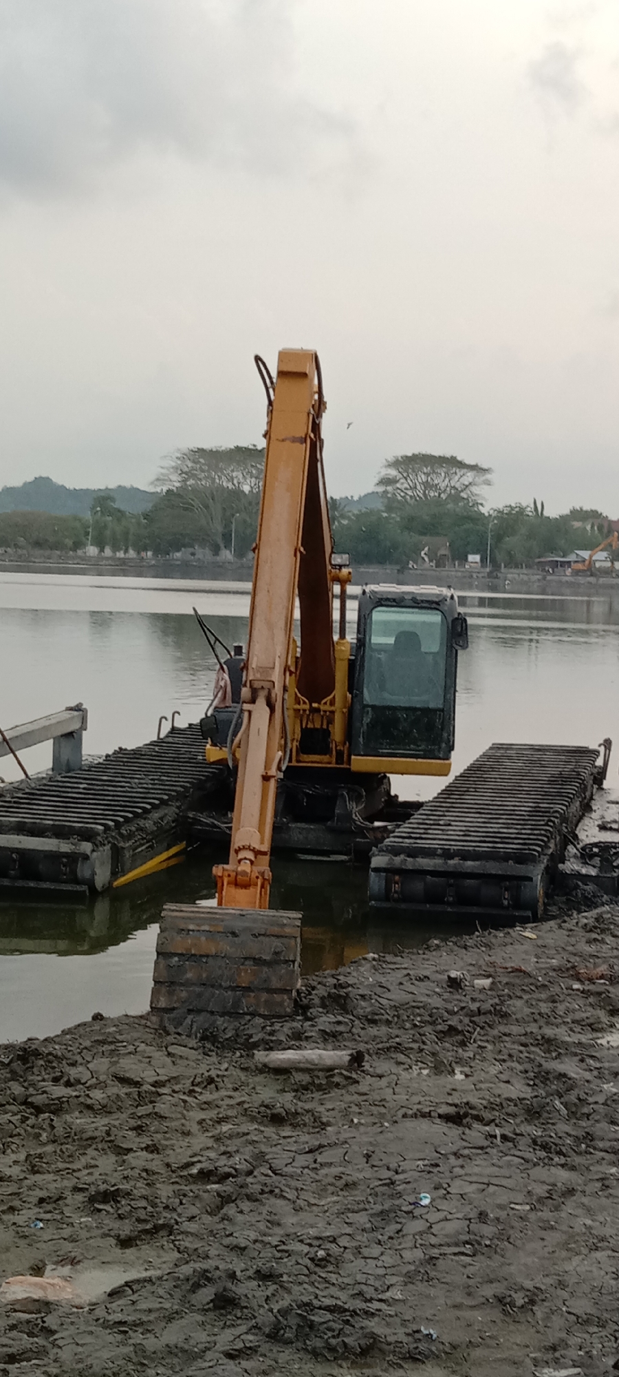 An orange amphibious excavator stands partially submerged in a body of water near a muddy bank. The excavator is fitted with two black, rectangular pontoons. Its arm is raised, and the bucket rests on the muddy shore. The water extends to the horizon, and in the distance, trees and a low-lying mountain range are visible. The sky is overcast. The location is in the city of Lhokseumawe, Indonesia.