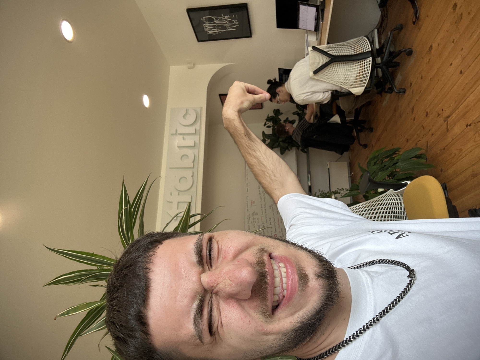A selfie captures a close-up of a man with dark hair and a beard, wearing a white t-shirt and a silver chain necklace. His face is contorted in a squinting smile, and he holds his arm up in the air. Behind him is an office setting. A sign with the word "fabric" is mounted on a wall. Another person sits at a desk working, in front of a computer. There are office chairs, plants and a wooden floor visible. A framed piece of art is mounted on the wall above the worker. The location is likely in Sofia, Bulgaria.