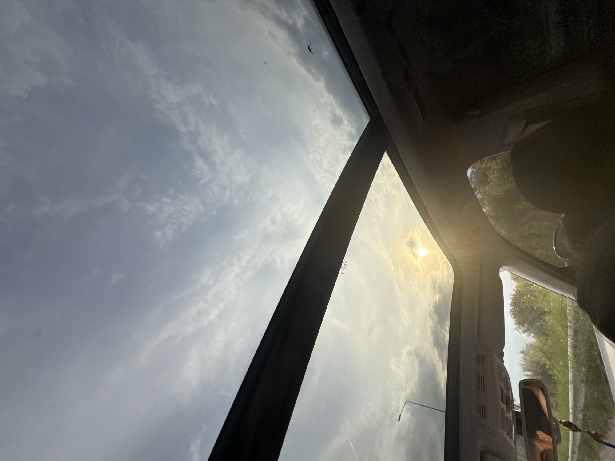 The image depicts an upward-angled view from the interior of a vehicle located in Suprunivka, Ukraine, looking through a panoramic sunroof toward the sky during late afternoon or early evening. The sky, partially visible through the large glass panel, exhibits a mixture of scattered cumulus and stratus clouds, illuminated by the low-hanging sun. The sun, positioned in the lower-right quadrant of the sunroof, emits a strong golden light and significant lens flare, creating a radiant glow across the lower portion of the sky. A prominent, dark structural bar of the sunroof horizontally bisects the frame. Below this bar, the interior of the vehicle is partially visible, including the headliner, overhead console, and a rearview mirror from which a colorful decorative charm hangs. The mirror's reflection is indistinct but shows a person's eyes or forehead. Outside the vehicle, on the left, a street light on a pole rises against the sky. Further down, green foliage, possibly trees and bushes lining a road, is discernible beyond a grey metal road barrier. Minor dust particles or imperfections are visible on the sunroof glass.