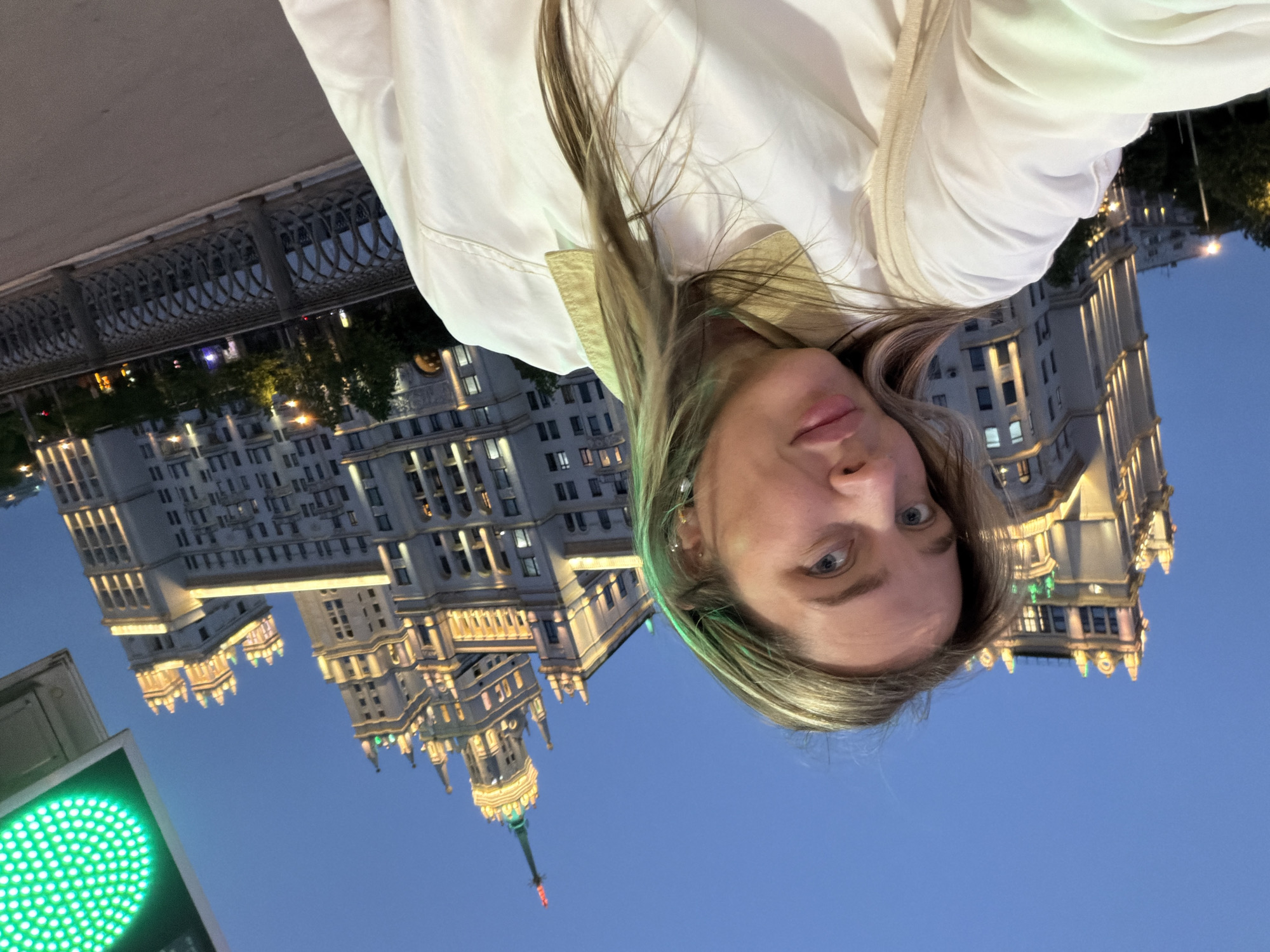 A vertical selfie depicts a fair-skinned woman with blonde hair in front of an illuminated building at dusk. Her eyes are open, and she is wearing a white jacket. In the lower left corner of the frame, there is a green traffic light. The building behind her features a spire and is lit up with bright white lights, contrasting against the blue sky. The location is likely urban with elaborate architecture. The image suggests a moment of personal documentation against an impressive cityscape. There is no indication the image was taken in Перовское сельское поселение, Ukraine.