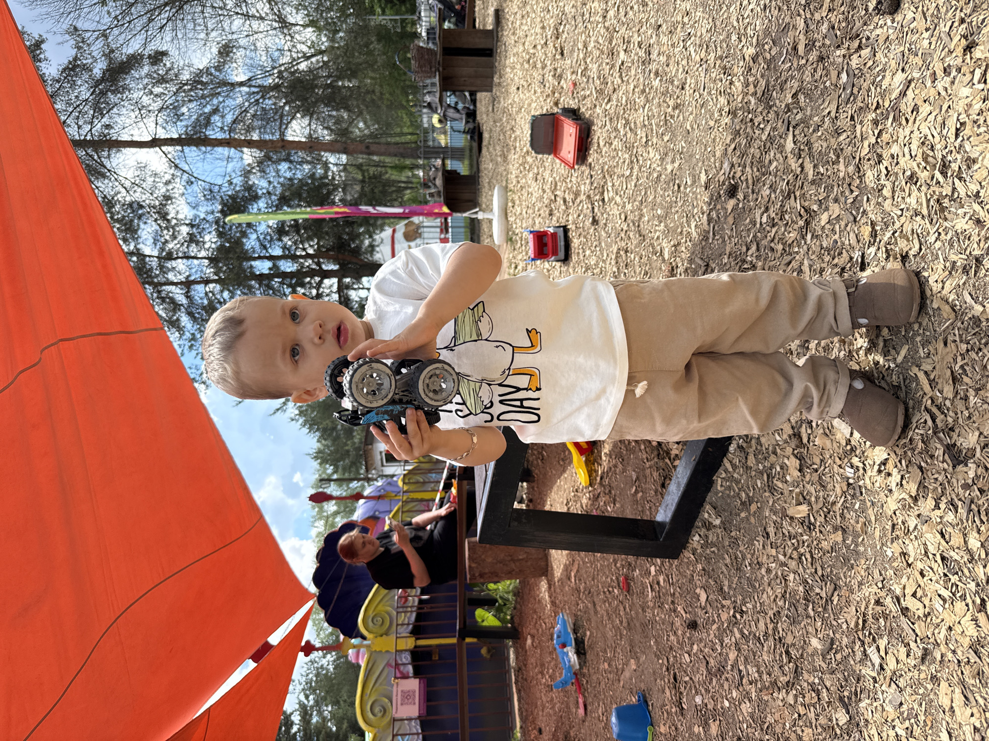This image captures a bright, daytime scene in an outdoor play area in Rusanovo, Russia. The primary subject is a young, light-haired boy, likely a toddler or preschooler, who is standing and looking directly at the camera with a neutral to curious expression. He is dressed in a white t-shirt featuring a cartoon duck graphic and the visible text "HAPPY DAY!", tan pants, and brown shoes. The boy holds a blue and black toy car in both hands, seemingly presenting it.
The ground of the play area is covered extensively with light-colored wood chips, a common feature in playgrounds. Scattered around the ground are various other toys, including a red toy truck, a blue toy truck, a blue bucket, and a blue water gun. To the boy's right, a black metal frame, possibly part of a bench or a play structure, extends into the foreground.
In the mid-background, partially obscured by the frame and a large, vivid orange shade canopy that covers much of the left side of the frame, a woman with red hair is seated on a bench or similar structure. She is wearing dark clothing and appears to be engrossed with a mobile device. The background consists of numerous trees, likely conifers, and patches of blue sky with white clouds, indicating clear weather. The scene conveys a relaxed and active atmosphere, characteristic of children playing outdoors.