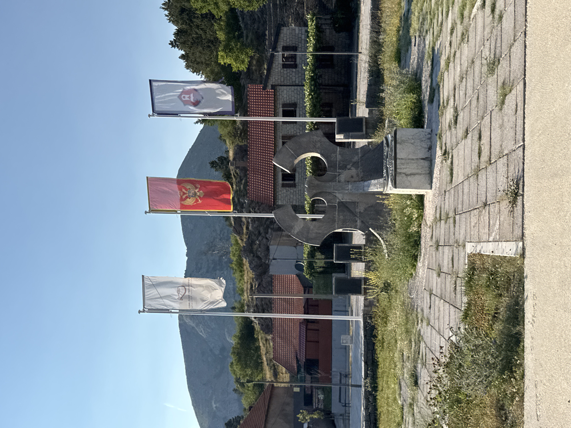 The image shows a monument in the city of Kopito, Montenegro. The monument features a large, abstract stone sculpture with two large curved elements meeting at the top, resting on a rectangular stone base. Three flags are flown on flagpoles to the right of the sculpture. The leftmost flag is white with an emblem in the center. The middle flag is red with a double-headed eagle emblem, which is the national flag of Montenegro. The rightmost flag is white with a blue border and an emblem of a red shield with a white crest. In the background, there are several stone buildings with red tile roofs, surrounded by vegetation and a rocky hillside with trees. The foreground shows a paved courtyard with square stone tiles, some of which are cracked and overgrown with grass. The sky is clear and blue, indicating a daytime scene. There are no people visible in the image.
