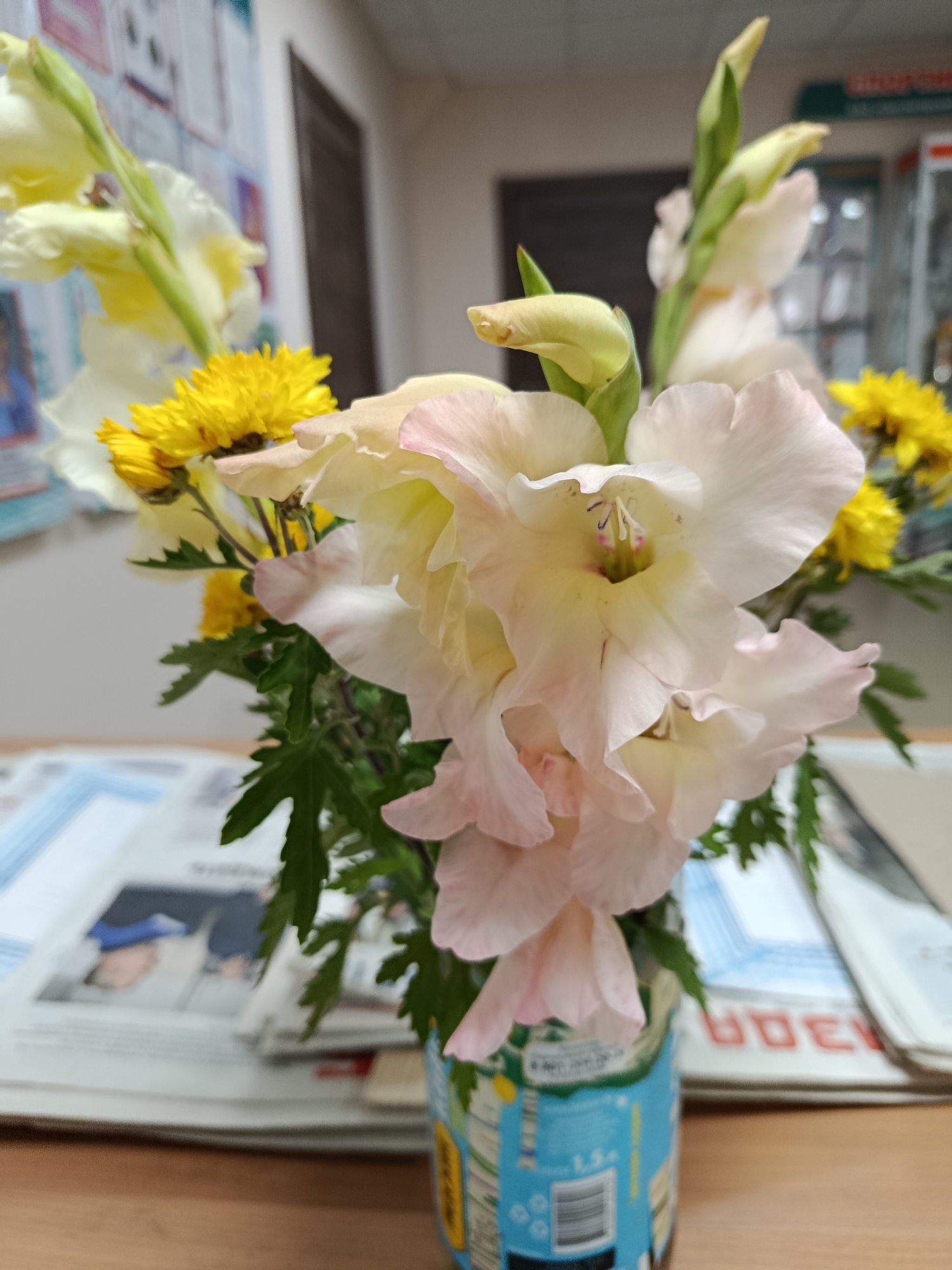 A bouquet of gladiolus flowers with delicate pink and white petals, interspersed with bright yellow chrysanthemum-like flowers, is placed in a blue and white patterned vase on a wooden desk. The vase appears to be made from a recycled container. Behind the flowers, several newspapers or magazines are stacked, with some visible text and images. The background is blurred, showing a wall with framed items and a doorway, suggesting an indoor setting, possibly an office or a room in a residential building in Svietlahorsk, Belarus. The lighting suggests it is daytime.