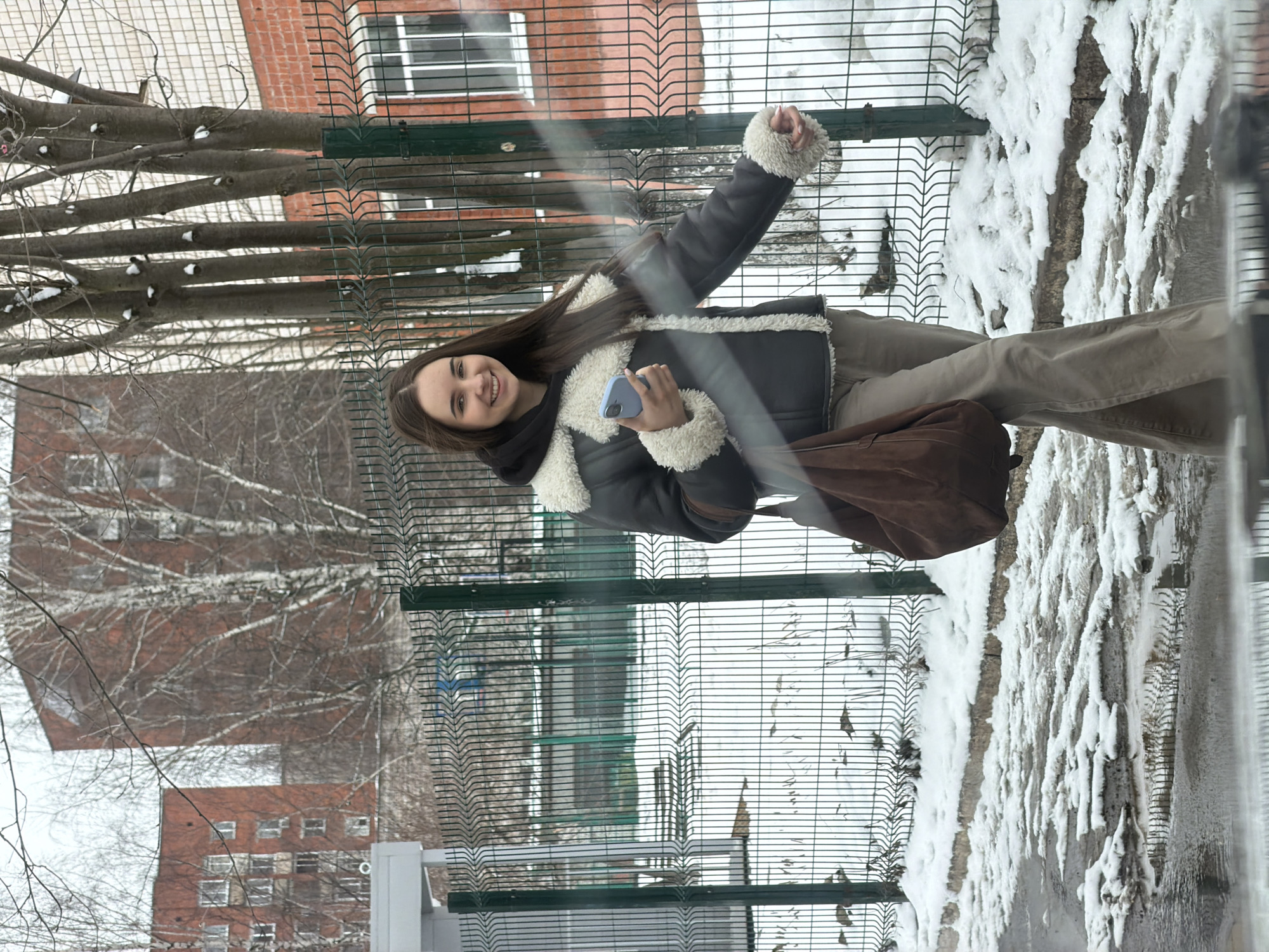 A young woman stands near a green metal fence with snow on the ground in Izhevsk, Russia. She is holding a blue smartphone, wearing a jacket with a white faux fur trim, khaki pants, and carrying a brown bag. She is smiling. Behind the fence, there is a brick building and bare trees. The scene is slightly distorted, as if viewed through a window.