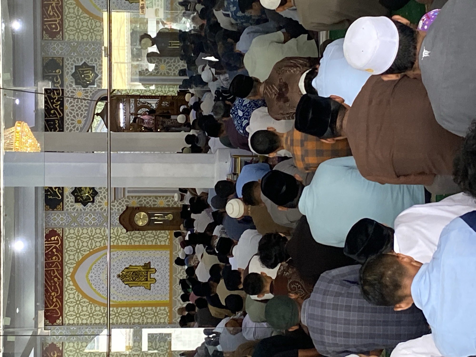 The image shows a gathering of men inside a mosque. The men are seen from behind, seated and facing forward, presumably during a prayer service. They are dressed in various attire, including traditional clothing and caps. The mosque interior features decorative walls with Islamic calligraphy and patterns, a grandfather clock, and a raised wooden pulpit where a person is standing and addressing the congregation. Sunlight streams in from windows, illuminating the space. The overall scene depicts a religious gathering in Lhokseumawe, Indonesia.