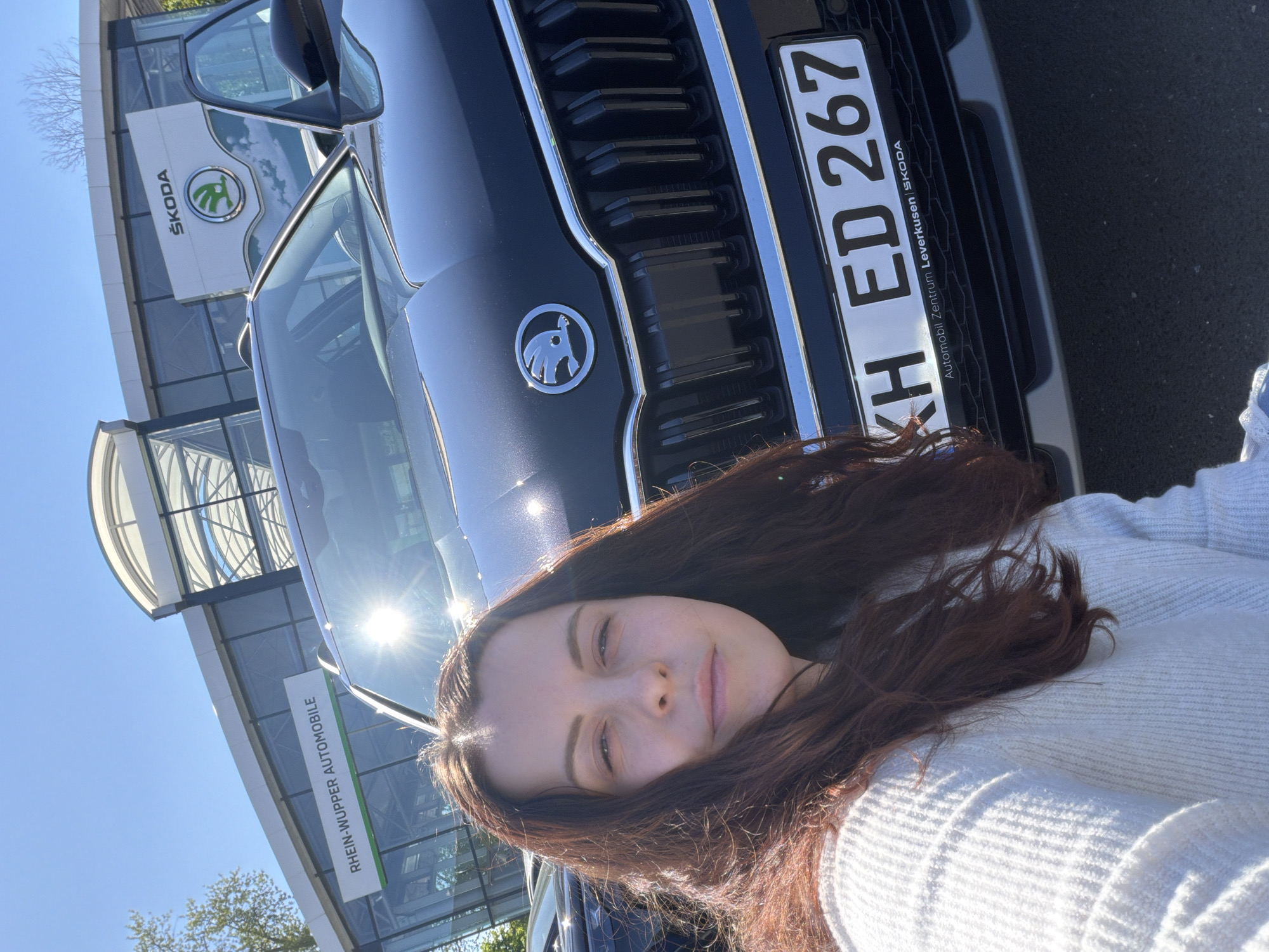 The image is a selfie of a woman in front of a dark-colored Škoda car. The car's license plate reads "KH ED 267" and includes the text "Automobil Zentrum Leverkusen ŠKODA", indicating the location is likely a Škoda dealership in Leverkusen, Germany. In the background, there is a building with the Škoda logo and the name "RHEIN-WUPPER AUTOMOBILE". The weather appears sunny. The woman is wearing a light-colored sweater.