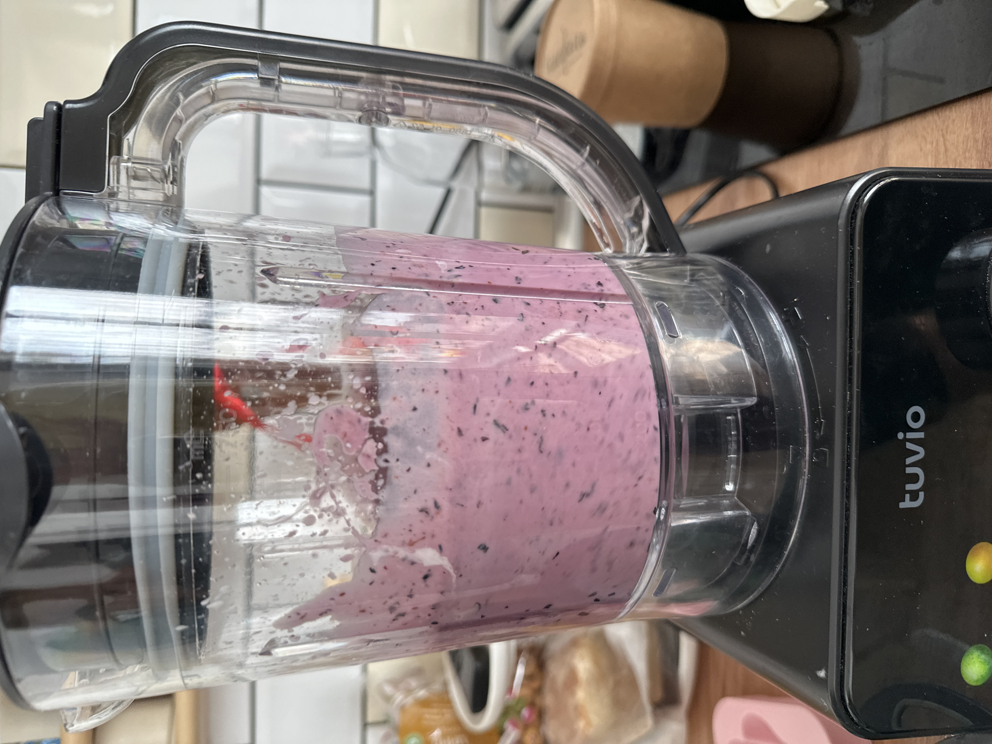 An eye-level, close-up shot shows a blender with a pinkish-purple smoothie being made. The blender is black with a clear plastic jug, and the smoothie is splashing upwards. In the background, a tiled wall and some kitchen counter items are visible. The base of the blender has a brand name "tuvio" printed on it, along with two illuminated circular buttons, one green and one yellow. The setting appears to be a kitchen in Vsevolozhsk, Russia.