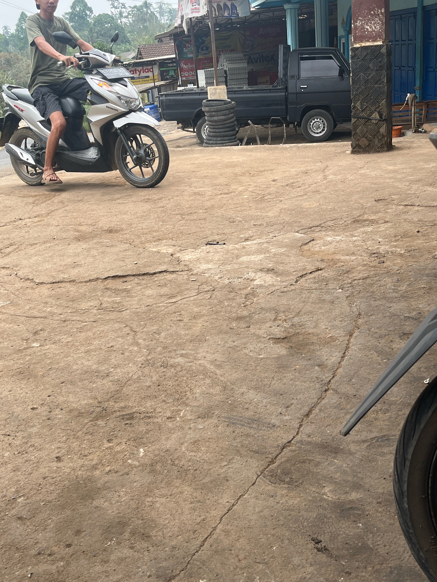 In Karang Sari, Indonesia, a young man wearing a green t-shirt and black shorts is seated on a white Honda motorcycle. He is wearing brown sandals. The motorcycle is parked on a cracked, dirt-covered concrete surface. In the background, a black pickup truck is visible, along with stacks of tires. Buildings with blue doors and tiled roofs are also present. The environment appears to be an outdoor area, possibly a roadside shop or workshop, with a slightly overcast sky.