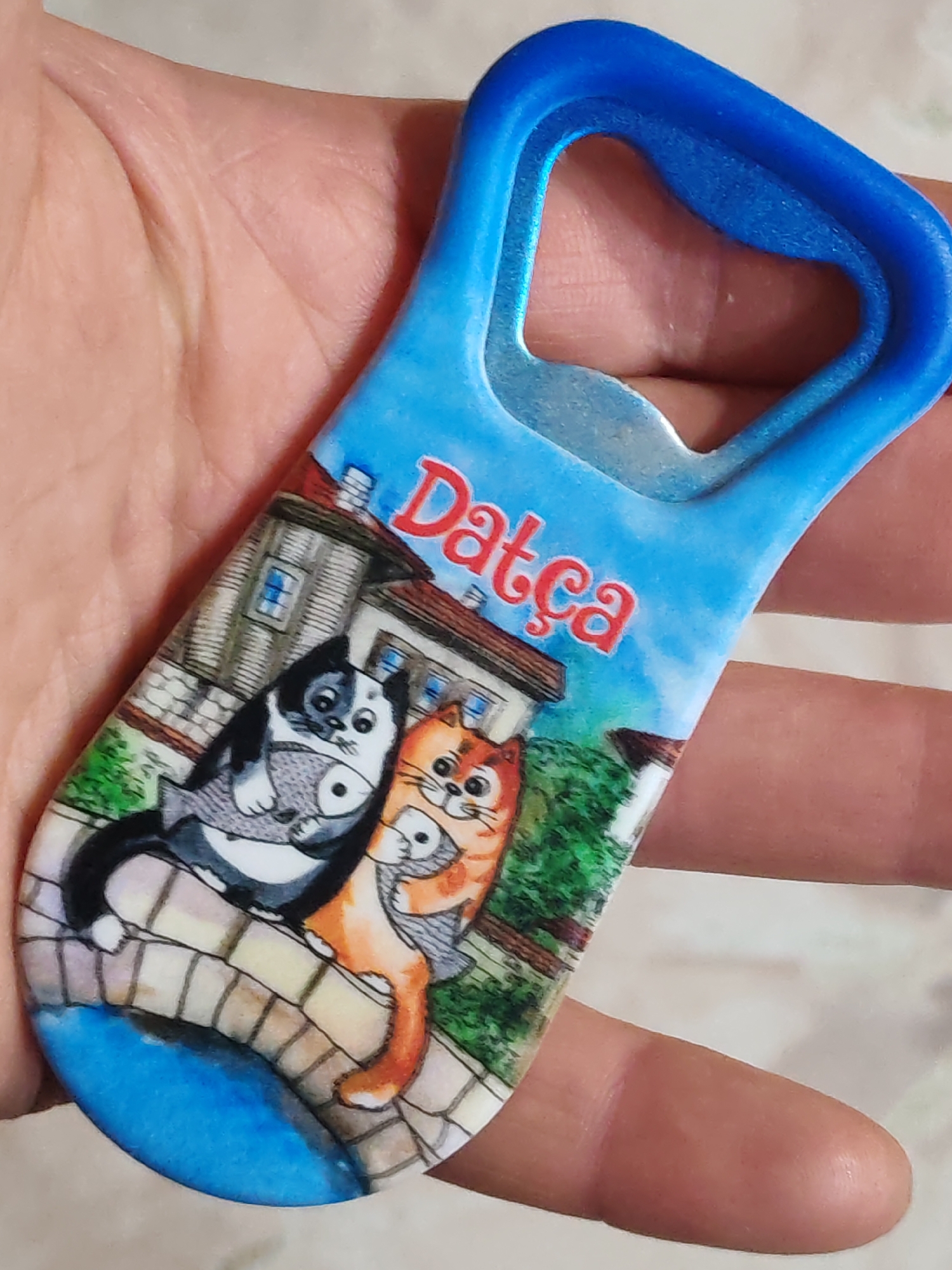 A hand is holding a souvenir bottle opener with a design depicting two cartoon cats on a stone bridge in Datça, Turkey. The bottle opener has a blue handle and a metal opener portion. One cat is black and white and holding a fish, while the other cat is orange. Buildings are visible in the background, and the word "Datça" is written in red above the cats. The lower portion of the bottle opener depicts a blue body of water under the bridge.