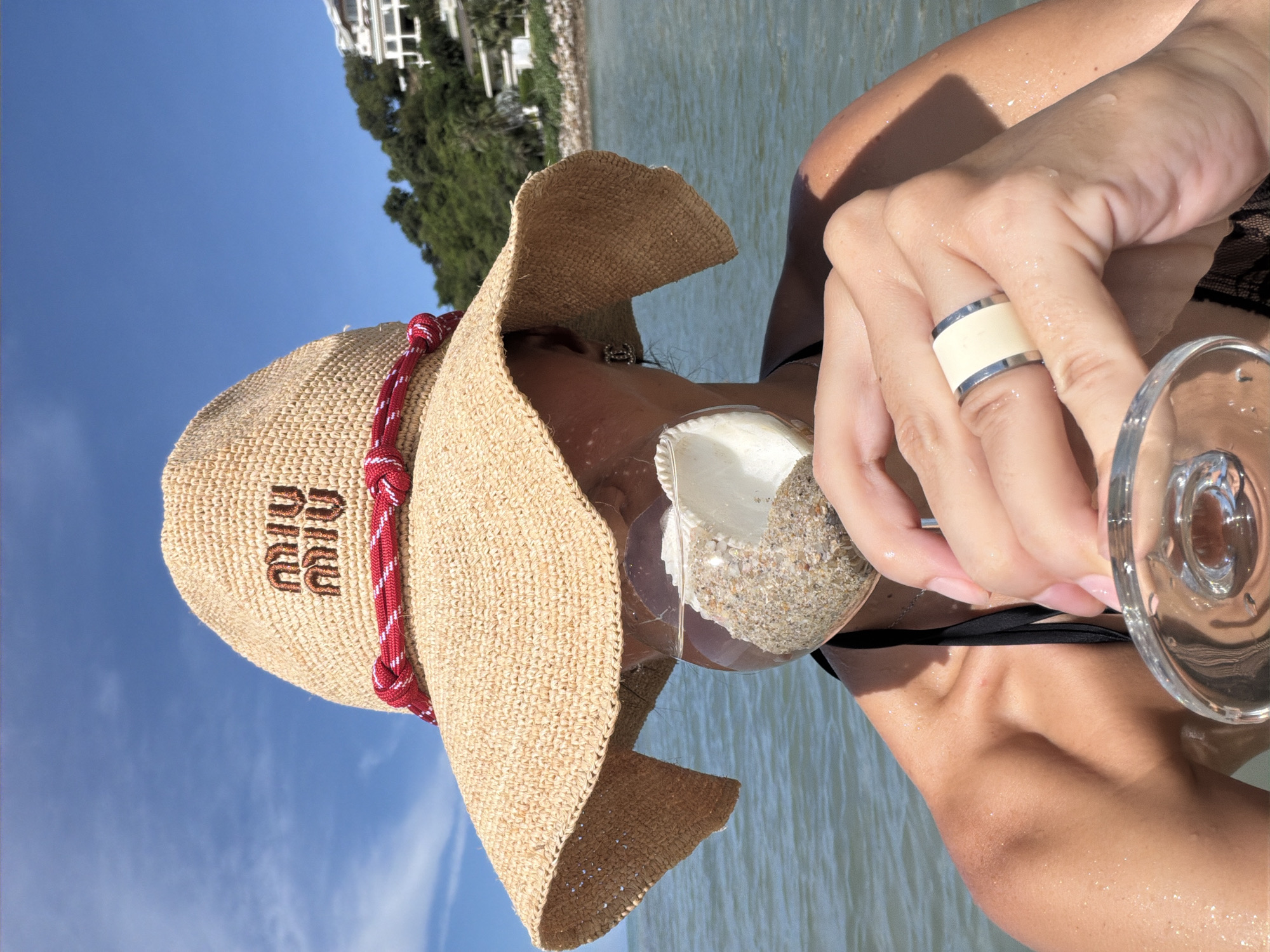 A person wearing a wide-brimmed straw hat with a red and white rope band is holding a clear glass with sand and a seashell inside. The hat has the logo "MIU MIU" embroidered in brown. The person is also wearing a ring with a cream-colored band and silver edges on their right hand. Their skin is wet, suggesting they are in or near water.
The scene appears to be a beach or seaside location, with calm water in the background and lush green trees and a building visible on a hillside. The sky is a clear blue with some faint clouds, indicating it is daytime with good weather. The lighting suggests it is a sunny day, possibly late morning or early afternoon. The overall mood is relaxed and possibly vacation-like. The person's face is partially obscured by the hat. The visible part of the person's attire is a black bikini top. The sand in the glass appears coarse and mixed with small shells. The image is oriented vertically.