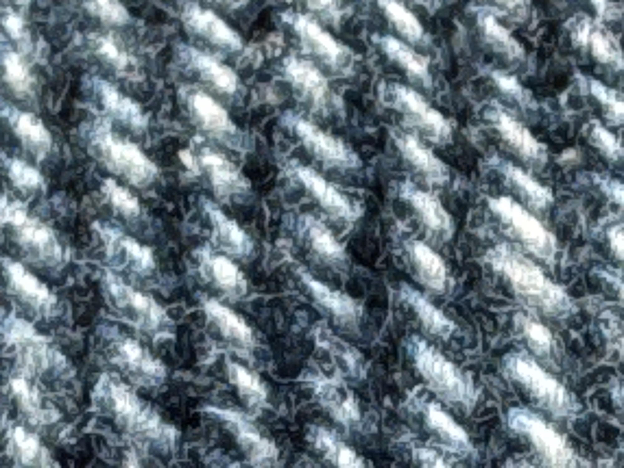 The image is a close-up, abstract view of a knitted or woven material, likely fabric. The texture is highly detailed, showing intertwined fibers that create a diagonal pattern of raised ridges and textured depressions. The colors are predominantly shades of dark gray, black, and white, with some hints of blue. The lighting creates highlights and shadows, emphasizing the three-dimensional nature of the weave. There are no discernible objects, people, or actions visible. The image offers no context related to a specific location, including Strümpfelbrunn, Germany.