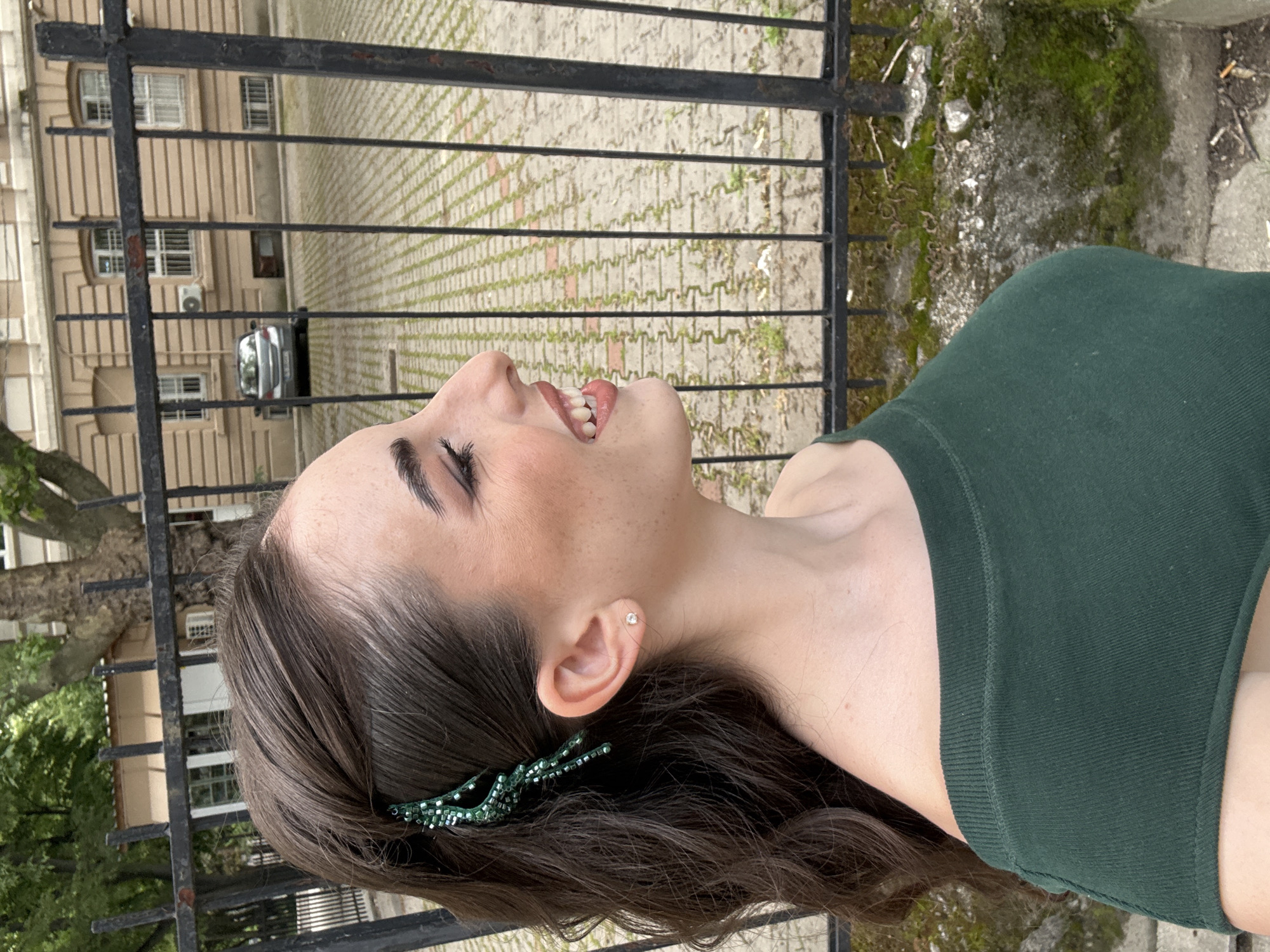 A young woman with brown hair and a green top is outdoors. She is smiling with her head tilted back, positioned in front of a black metal fence. Through the fence, a building with multiple windows and a silver car can be seen. The background also features a brick pathway and some greenery. A green hair accessory is visible in her hair, near her ear. The setting appears to be a city street, plausibly in Plovdiv, Bulgaria, although visual confirmation of specific landmarks is absent.