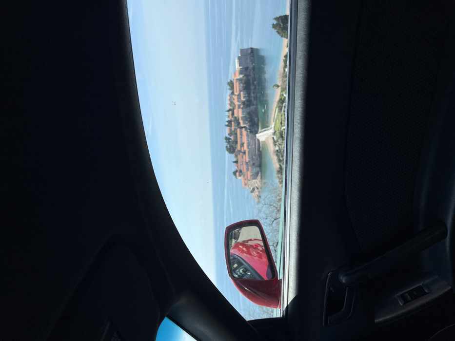 The image displays a static outdoor scene, captured from within a vehicle looking through its side window. The top section of the frame shows the vehicle's dark interior headliner and A-pillar. The bottom right area reveals a dark door panel equipped with a handle and window control buttons. A red exterior side mirror is positioned on the left, reflecting a portion of a red vehicle's body.
The primary subject outside the vehicle is the island of Sveti Stefan, located in Montenegro, situated in calm, blue-green seawater under a clear, bright blue sky. The island is densely covered with numerous stone buildings featuring orange terracotta roofs, collectively forming a fortified appearance. A narrow, light-colored causeway extends from the island, connecting it to the mainland. The adjacent mainland shoreline exhibits sparse, dry vegetation and rocky terrain. No individuals, overt actions, or interactions are visible within the depicted scene.