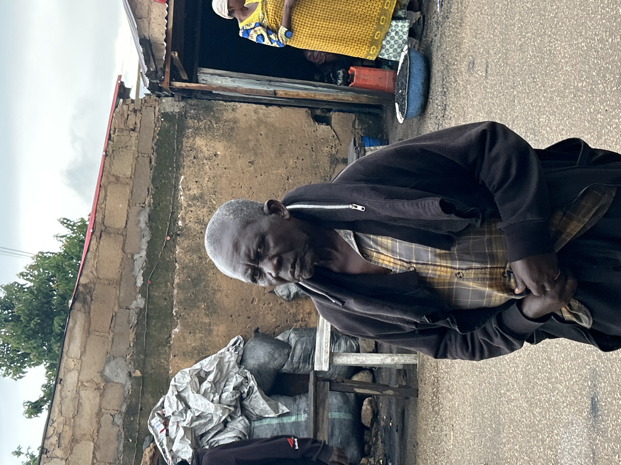 The image depicts an outdoor scene in the city of Jos, Nigeria, likely in a market or roadside area. In the foreground, an elderly Black man stands facing the camera. He has short, graying hair and is wearing a plaid collared shirt under a dark zip-up jacket. The jacket is draped loosely around his shoulders. His hands are clasped in front of him, and he wears a ring on his left hand.
Behind him, to the left, are several large sacks, some appearing to be covered in white material, suggesting merchandise or goods. A wooden table or stand is visible in front of these sacks.
To the right, a doorway leads into a shaded structure. A woman, also Black, is partially visible in the doorway, wearing a bright yellow and blue patterned dress and a head covering. She appears to be interacting with someone inside the structure, though the details are unclear. In front of the doorway, on the ground, is a blue basin filled with dark material, possibly coal or charcoal, and a red container.
The background consists of rough, unfinished brick buildings with corrugated metal roofs, characteristic of some urban structures in Jos. The ground is paved with asphalt or similar material. The sky is overcast. The overall impression is of a humble, working-class environment.