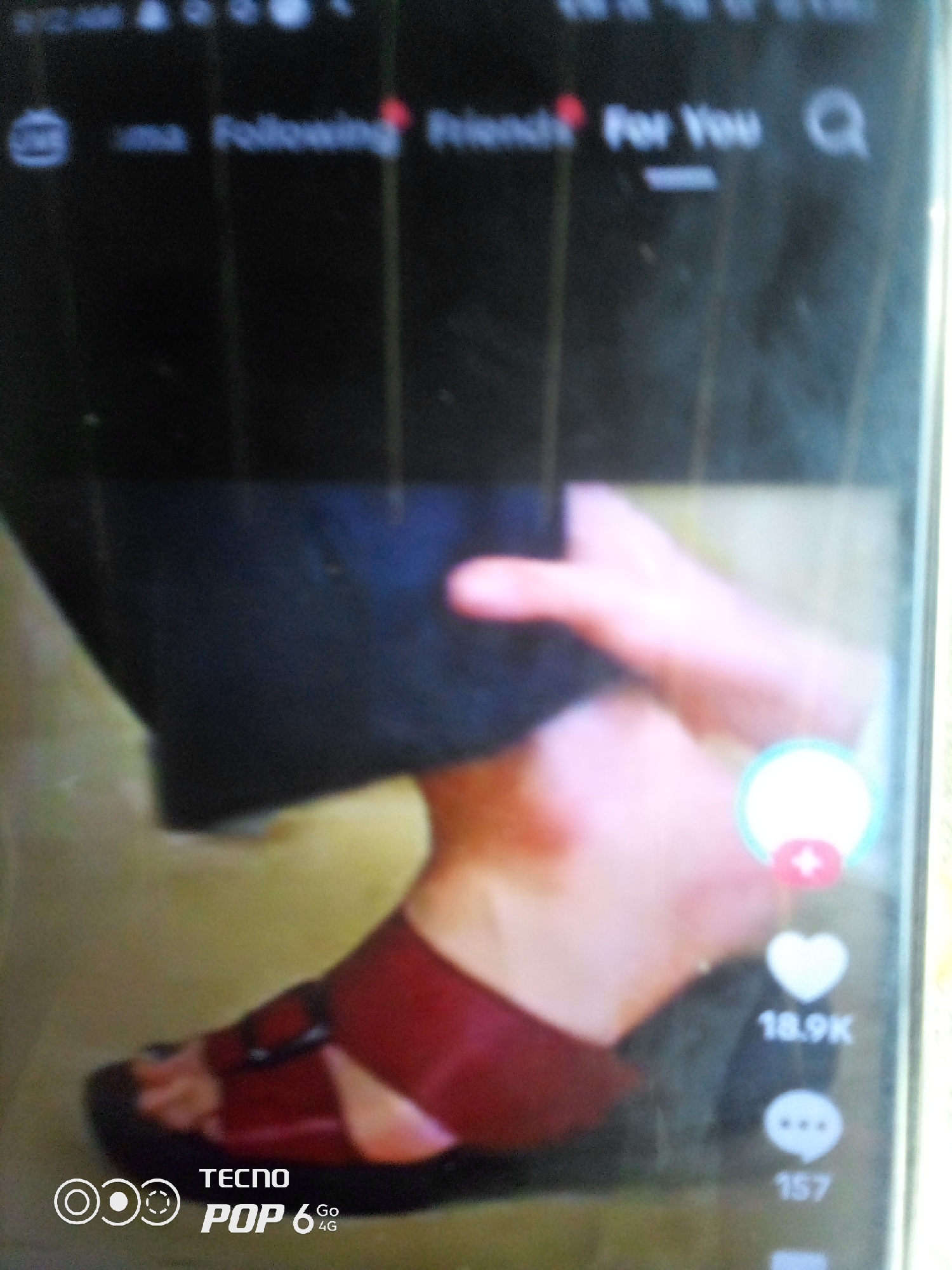 The image shows a close-up of a person's foot wearing a dark red sandal with a buckle. The sandal is placed on a textured surface that appears to be a floor. The background is blurred, but suggests an indoor setting. In the upper part of the image, there is a blurry screen interface, possibly from a social media app like TikTok, with text that reads "Following," "Friends," and "For You." There is also a search icon. On the right side of the foot, there are icons indicating engagement metrics, with "18.9K" likely representing likes and "157" possibly representing comments. At the bottom left of the image, there is a logo that reads "TECNO POP 6 4G." The overall impression is that this is a screenshot of a video or image from a social media platform, likely taken on a Tecno Pop 6 4G phone. The lighting suggests it was taken during the day.
