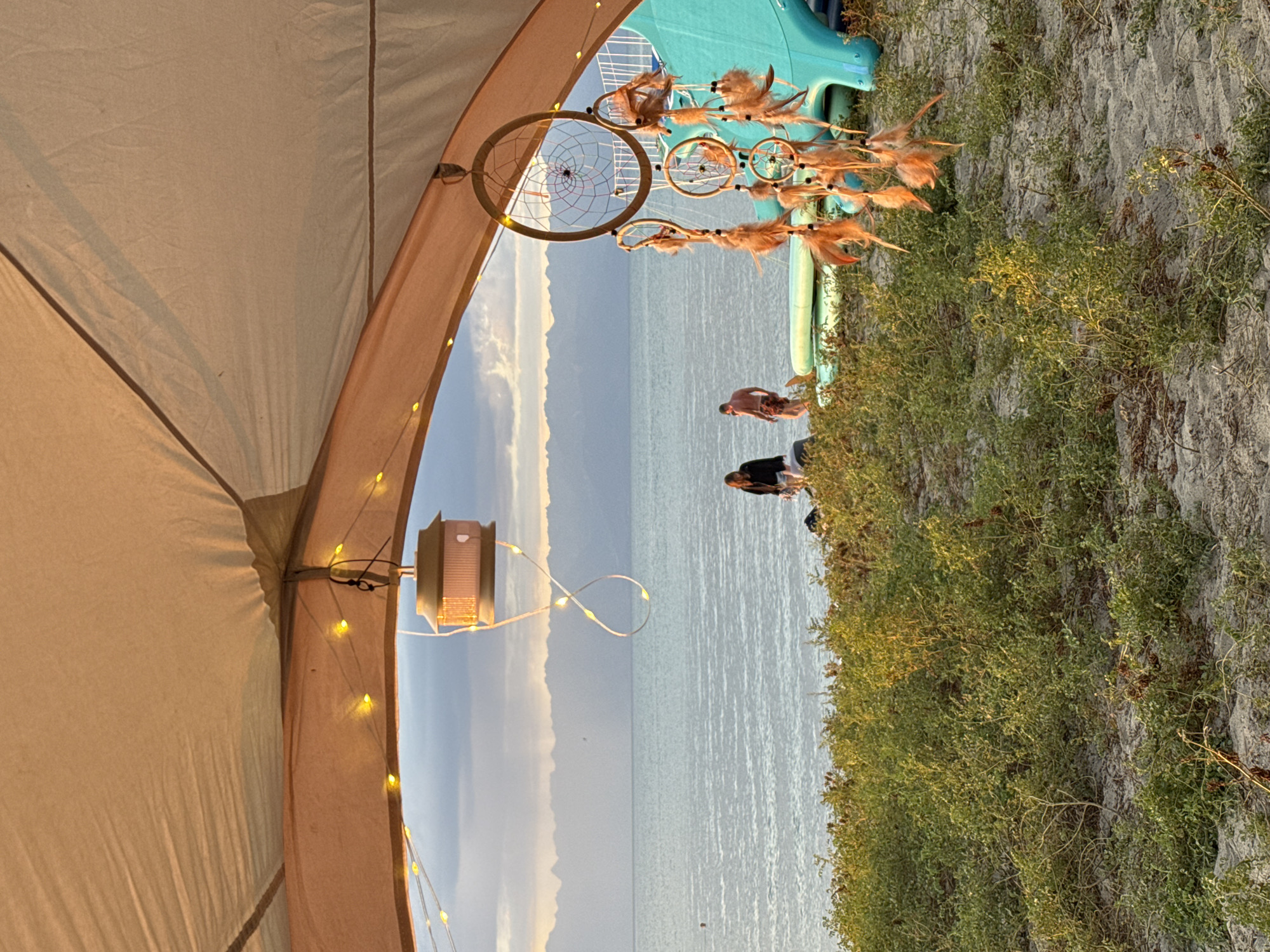 The image depicts a view from inside a tent towards a body of water, likely Lake Issyk-Kul, given the mountains in the background that are characteristic of the Issyk-Kul region near the city of Balykchy, Kyrgyzstan. The foreground features a sand-covered ground with low-lying green vegetation. A string of yellow fairy lights is draped across the tent opening, illuminating a hanging solar-powered lantern. To the right, a dreamcatcher with feathers is suspended from the tent. In the middle ground, two individuals are visible near the water's edge. One person, a woman with dark hair, is standing, while a man with a bare torso and patterned shorts is walking. They are near several inflatable watercrafts, including what appears to be an inflatable water slide. The water is calm with ripples reflecting the soft, diffused light of what appears to be either dawn or dusk. The distant background is dominated by a range of hazy, blue mountains under a sky with clouds.