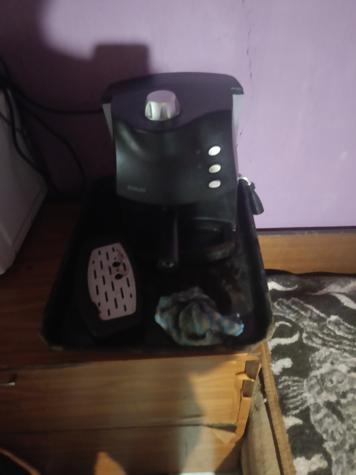 The image depicts a close-up, low-angle view of a black coffee maker sitting on a dark tray. The coffee maker has a light-colored dial on top and three white buttons arranged vertically on its front. To the left of the coffee maker, on the tray, is a light-colored drip tray with a slotted pattern and two circular indentations. A crumpled cloth, appearing to be blue and white patterned, is placed on the tray near the drip tray. The coffee maker and tray are positioned on a wooden surface, possibly a shelf or a table, which has a patterned rug underneath it. The background features a purple-tinted wall with shadows cast upon it. To the far left, a sliver of a white object, possibly an appliance, is visible. The scene appears to be indoors, in a residential setting, possibly in the city of Mizia, Bulgaria, though there are no definitive landmarks or indicators to confirm this specific location.