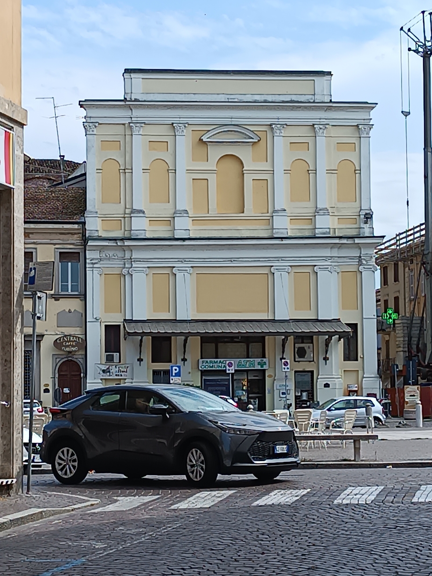 The image depicts a street scene in Casalmaggiore, Italy, featuring a dark gray hatchback car in the foreground, positioned on a cobblestone street with a pedestrian crossing. The car is angled towards the left, with its front facing forward. Behind the car, a light yellow building with classical architectural elements dominates the background. This building has a multi-story facade with columns, arched windows, and a prominent cornice. A sign that reads "FARMACIA COMUNALE" and "AFM" is visible on the ground floor, indicating a pharmacy. Several outdoor tables and chairs are set up on the sidewalk in front of the building. To the left of the main building, another building with a reddish-tiled roof is partially visible. In the distance, to the right, scaffolding and construction elements suggest ongoing work on another structure. The sky above is partly cloudy. There are no visible people interacting in the frame.