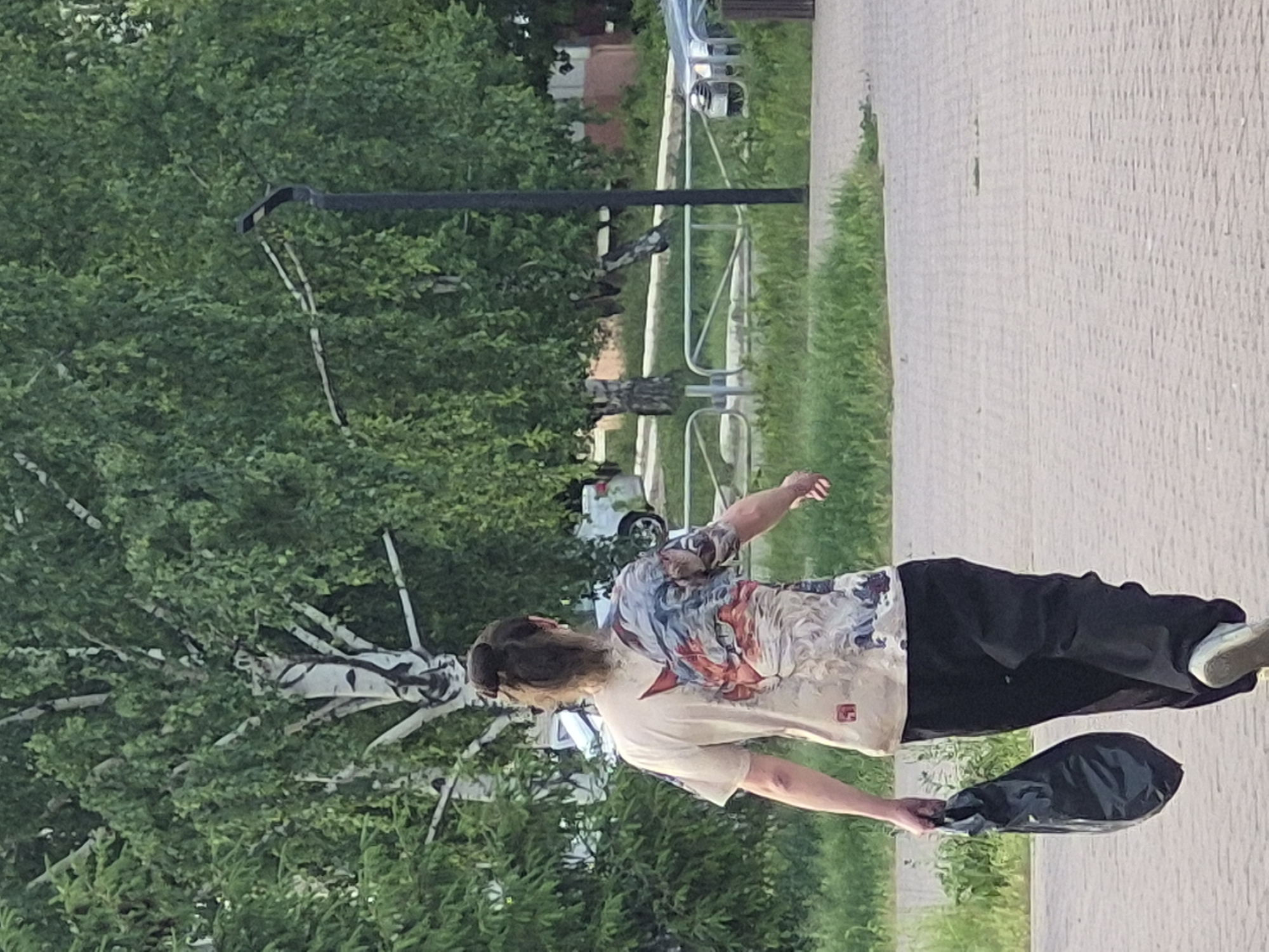 A person is walking on a paved sidewalk in an urban environment. The person is wearing a patterned t-shirt and dark pants, and is carrying a dark, possibly soft bag. The sidewalk is made of light-colored paving stones. Behind the sidewalk is a strip of green grass, and beyond that, there are trees and bushes. A metal barrier is visible behind the grass. A light-colored car is also visible in the background, partially obscured by the trees. A lamp post is seen over the trees.