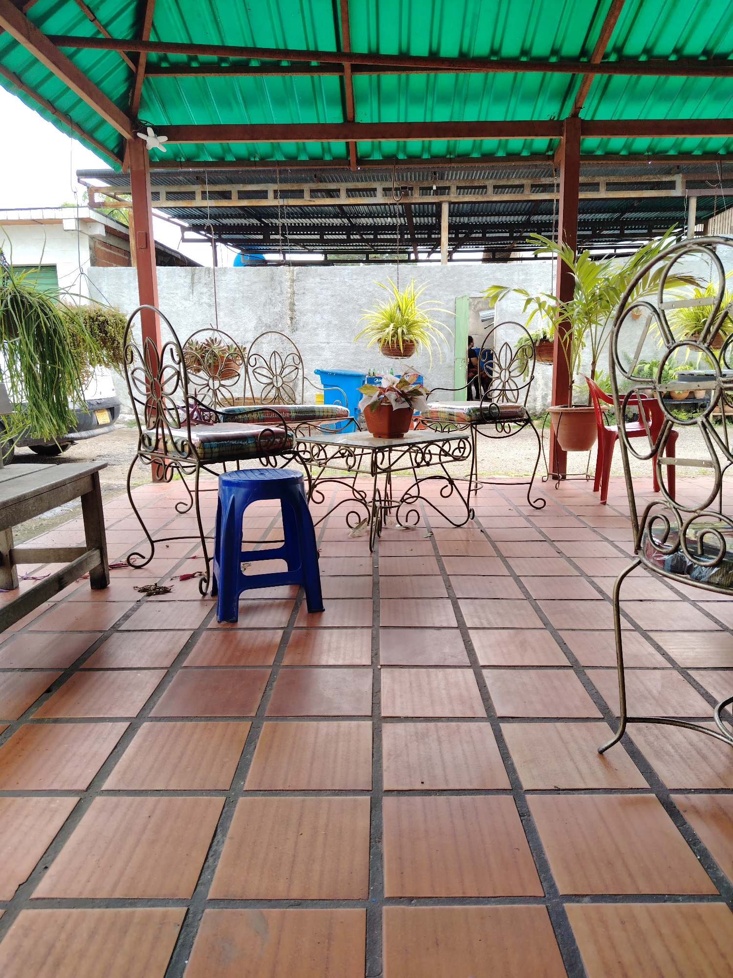 The image depicts an outdoor patio area in Tinaquillo, Venezuela, characterized by terracotta-colored tiled flooring and a green corrugated metal roof supported by brown beams. The patio furniture consists of ornate metal chairs with flower-shaped backs and cushioned seats, arranged around a matching metal table topped with a potted plant. A blue plastic stool is positioned in front of the table. Additional potted plants, some hanging, are visible. In the background, a white wall is present, along with glimpses of a building and vegetation. The setting appears to be a semi-enclosed space, possibly a covered terrace or veranda.