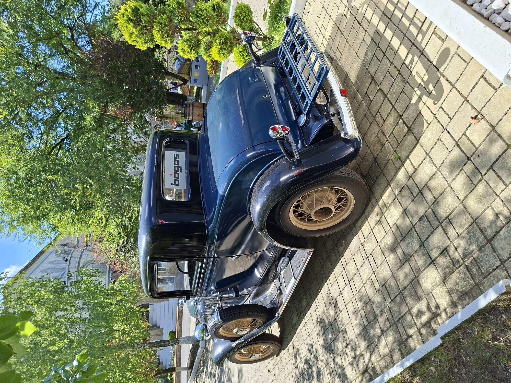 The image shows a dark blue vintage coupe parked on a paved area in front of a building, with trees and greenery surrounding it. The car, a classic model likely from the 1930s, features a spare tire mounted on the side, chrome accents, and a luggage rack at the rear. A logo with the text "BOGOS" and a lightning bolt symbol is visible on the rear window. The building in the background appears to be an older structure with multiple levels and arched windows. The ground is paved with light-colored rectangular stones, and shadows from the trees are cast across the surface. The environment suggests an outdoor setting, possibly a courtyard or a street in a town. Based on the location information, this scene is situated in Milestii Mici, Moldova. There are no people or active interactions depicted in the image.