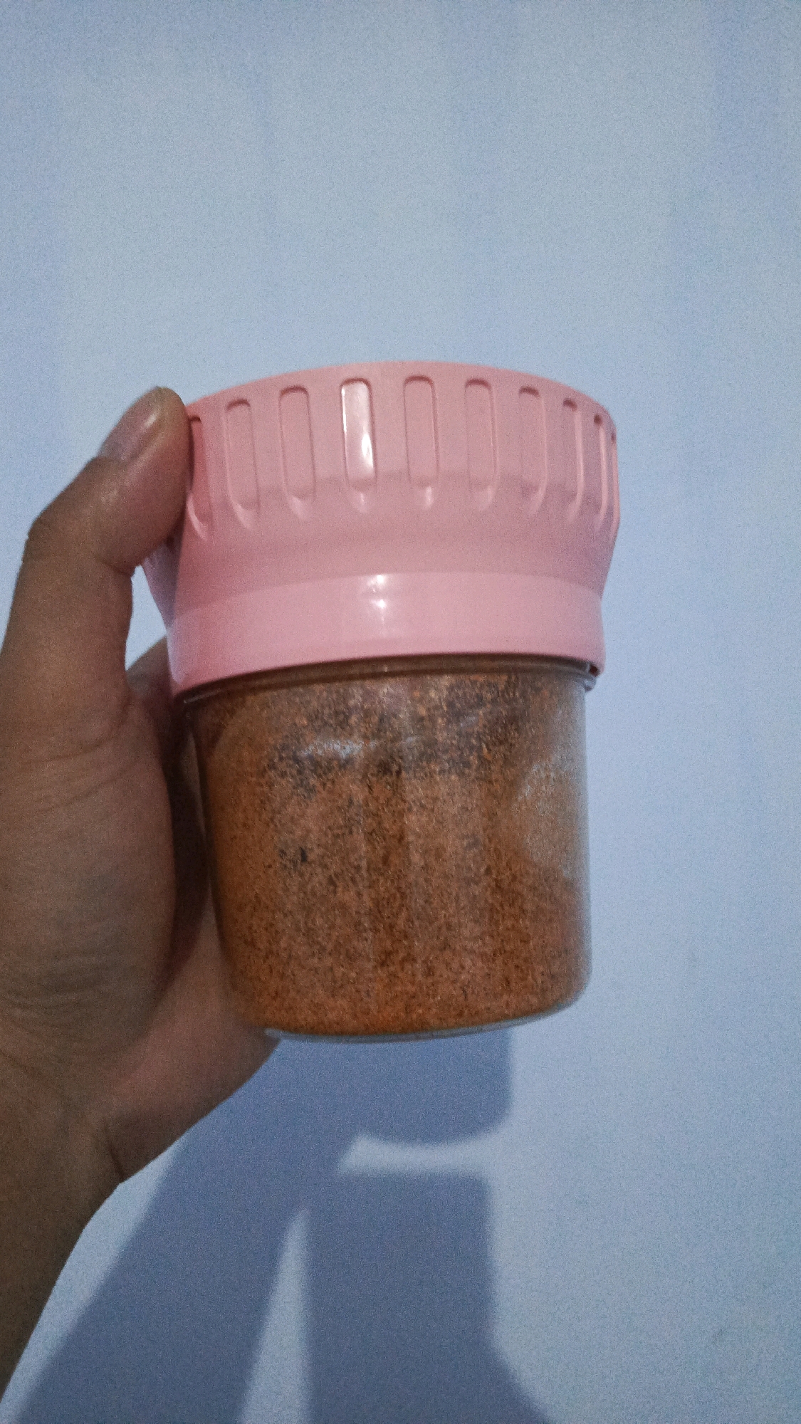 In Babadan, Indonesia, a person's hand is holding a transparent plastic container filled with an orange-brown granular substance. The container has a pink lid with a textured, ribbed design around its circumference. The background is a plain, pale blue wall.