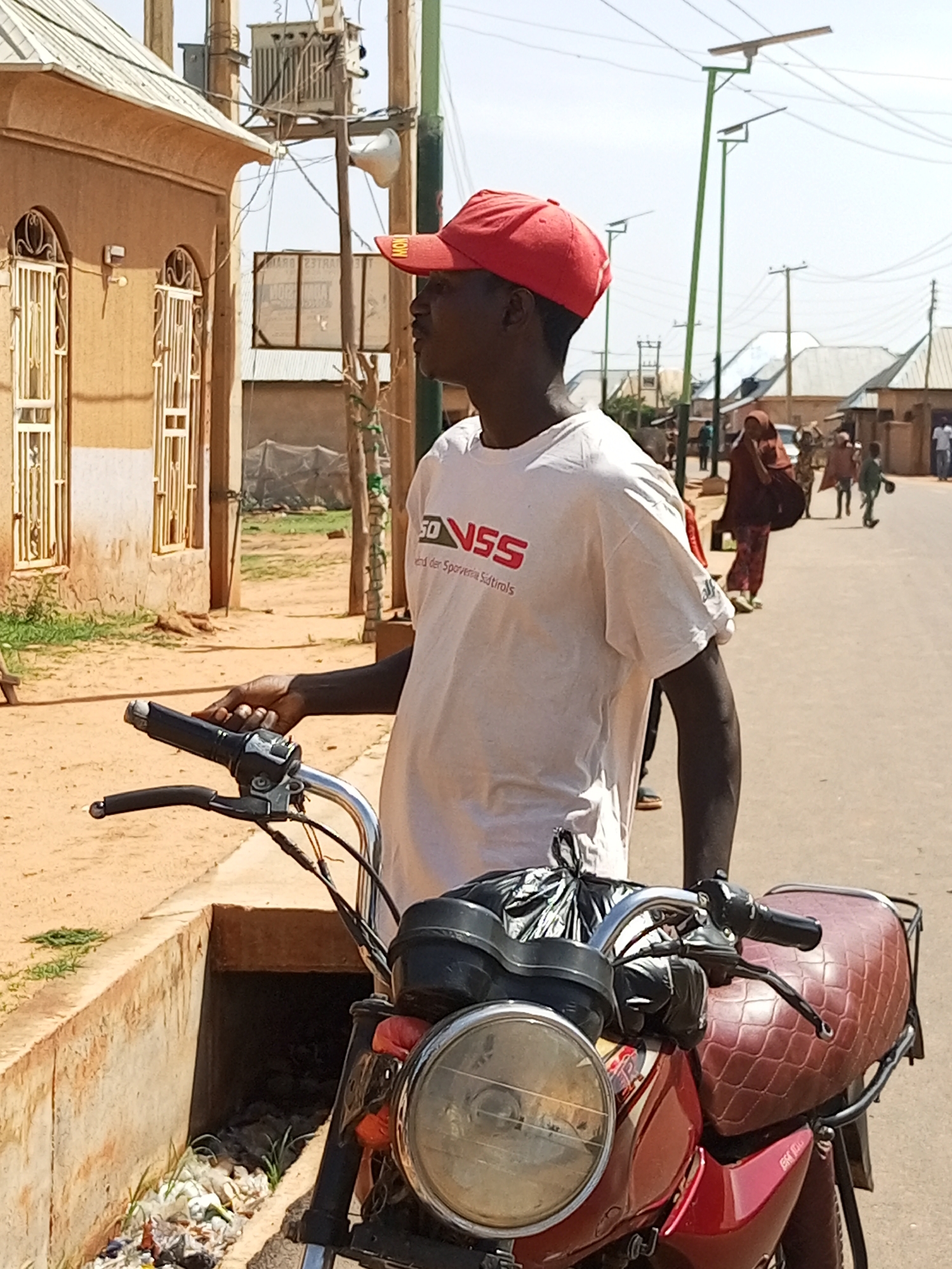 A young Black male wearing a red baseball cap and a white t-shirt with red text is standing next to a red motorcycle in Gombe, Nigeria. He is looking to his left, and his left hand is on the motorcycle's handlebar. The t-shirt has a logo with "VSS" in red and white, and text below it in German. The motorcycle has a quilted brown seat and a black plastic bag attached to the handlebars. Behind the man, there is a street with several people walking, some wearing traditional Islamic clothing. Buildings with light brown walls and arched windows with metal grates are visible to the left. There are power lines and streetlights in the background, consistent with an urban environment in Gombe. The ground is dusty and appears to be made of dirt. A concrete drainage ditch filled with trash and vegetation is located to the left of the motorcycle. The scene is illuminated by bright sunlight, suggesting it is daytime.