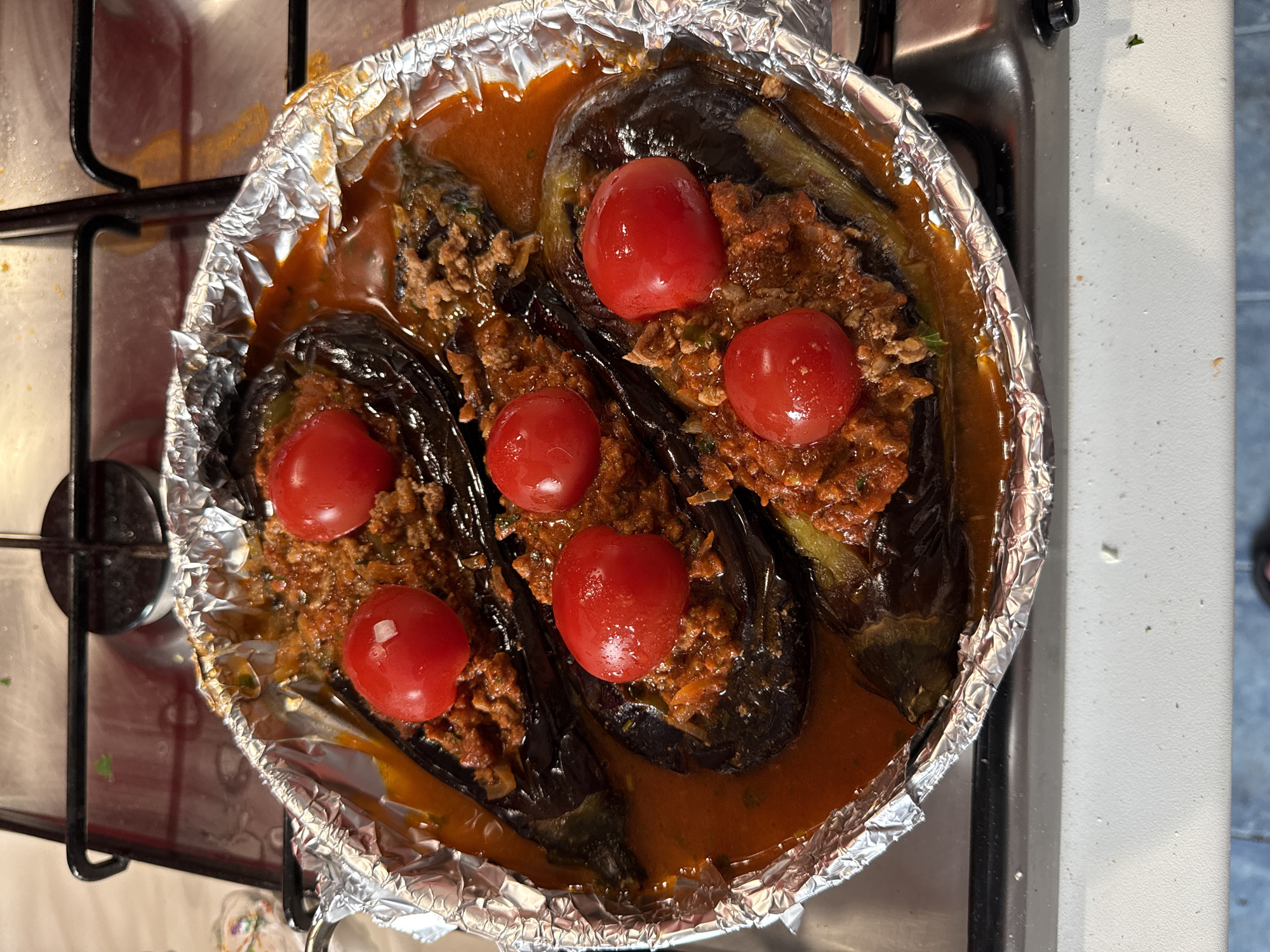The image shows a tray of stuffed eggplant with tomatoes, likely Imam bayildi or a similar dish, on a stovetop. The tray is lined with aluminum foil. The aubergines are stuffed with a meat-based mixture and topped with several red cherry tomatoes. A reddish-brown sauce surrounds the eggplants. The stove appears to be in a domestic kitchen setting. The image does not provide enough information to definitively identify the location as Milan, Italy.