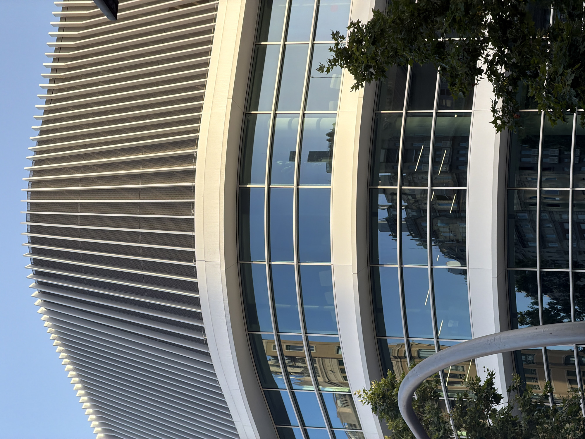 The image shows the contemporary facade of a building in Barcelona, Spain, featuring a curved structure with vertical silver fins on its upper section. The lower portion of the building is characterized by large, reflective glass windows that mirror the surrounding urban environment, including older, ornate architectural styles of adjacent buildings and trees. A metallic, curved sculpture is partially visible in the foreground, to the left of the frame, with a lush green tree on the right, obscuring a portion of the building. The sky is clear and blue, suggesting a sunny day. The overall impression is one of modern architecture set against a historic urban backdrop.