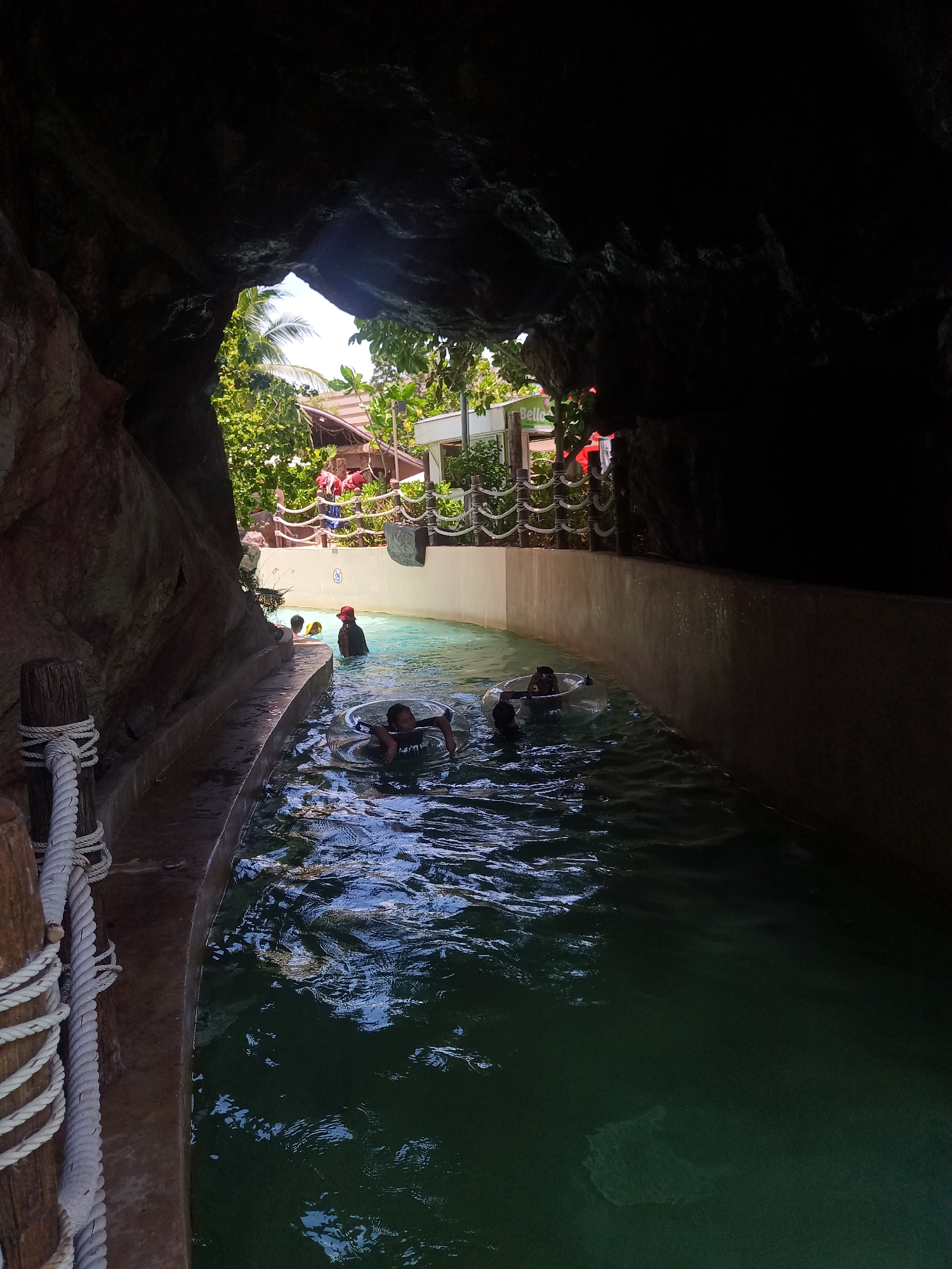 The photograph depicts a segment of a lazy river ride at a water park, possibly located in the Hua Hin City Municipality, Thailand, judging by the architectural style and vegetation visible in the background. The viewpoint is from within a faux rock tunnel section of the ride. The water is slightly murky and greenish-blue.
Several individuals are present in the water, some riding in inflatable inner tubes. One person is wearing a red hat. The water channel is bordered by a concrete wall. A wooden post with a rope attached is visible on the left side of the frame. In the background, beyond the tunnel opening, there's evidence of a resort or park area with buildings, decorative rope barriers, and trees. The lighting is a mix of bright sunlight in the exterior area and shadow within the tunnel.