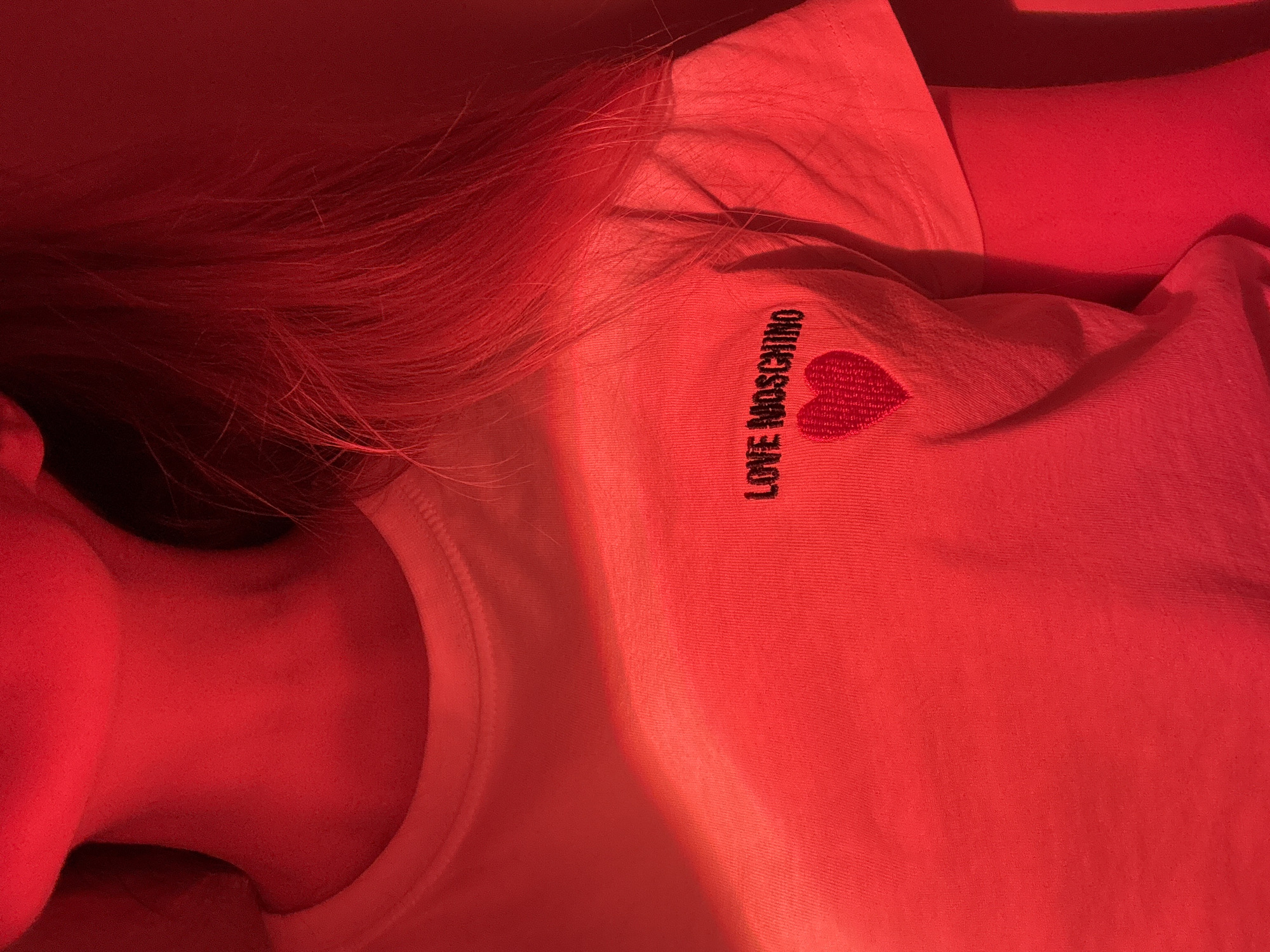 The image is a close-up of a person wearing a white t-shirt with a "LOVE MOSCHINO" logo and a heart emblem. The person is lying down, with their head and neck visible on the left side of the frame. The entire scene is illuminated with a red light, giving everything a red hue. The background is out of focus. Location information is not discernable from the image.