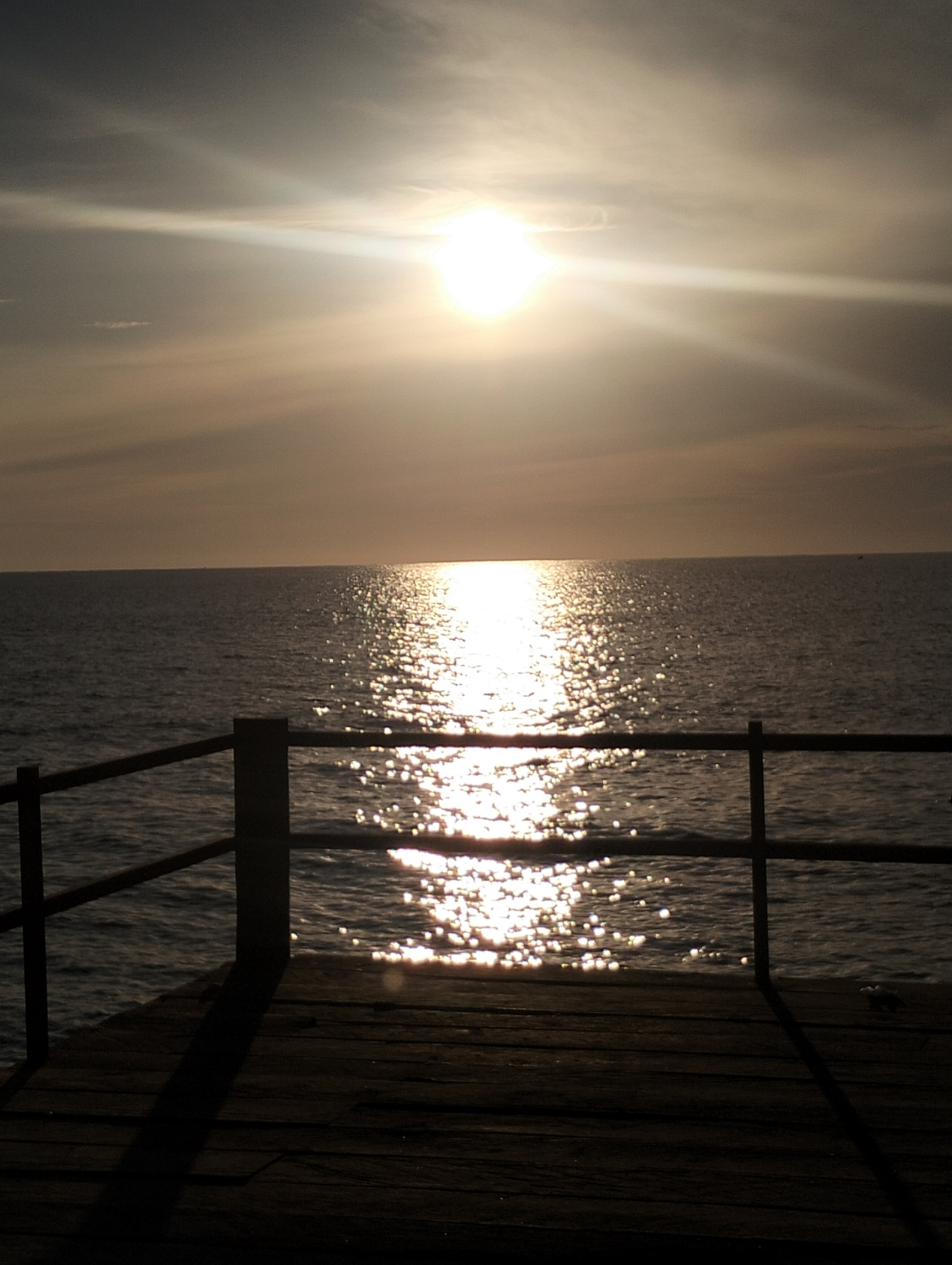 A wide-angle, vertical, low-angle shot shows a wooden dock and railing in the foreground, overlooking a body of water under a bright, hazy sun. The sun, high in the sky, casts a brilliant glare, reflecting off the water in a shimmering path that extends towards the horizon. The water itself appears calm, with gentle ripples visible. The sky is a pale gray, with wispy clouds that are partially obscured by the sun's brilliance. The wooden dock is dark and weathered, with visible planks and shadows cast by the railing. The railing consists of dark vertical posts and horizontal bars. In the far distance, the horizon is barely discernible where the water meets the sky. The scene evokes a sense of tranquility and a warm, late afternoon or early morning atmosphere. The image does not contain any discernible people or specific landmarks that would indicate its location within Lhokseumawe, Indonesia.