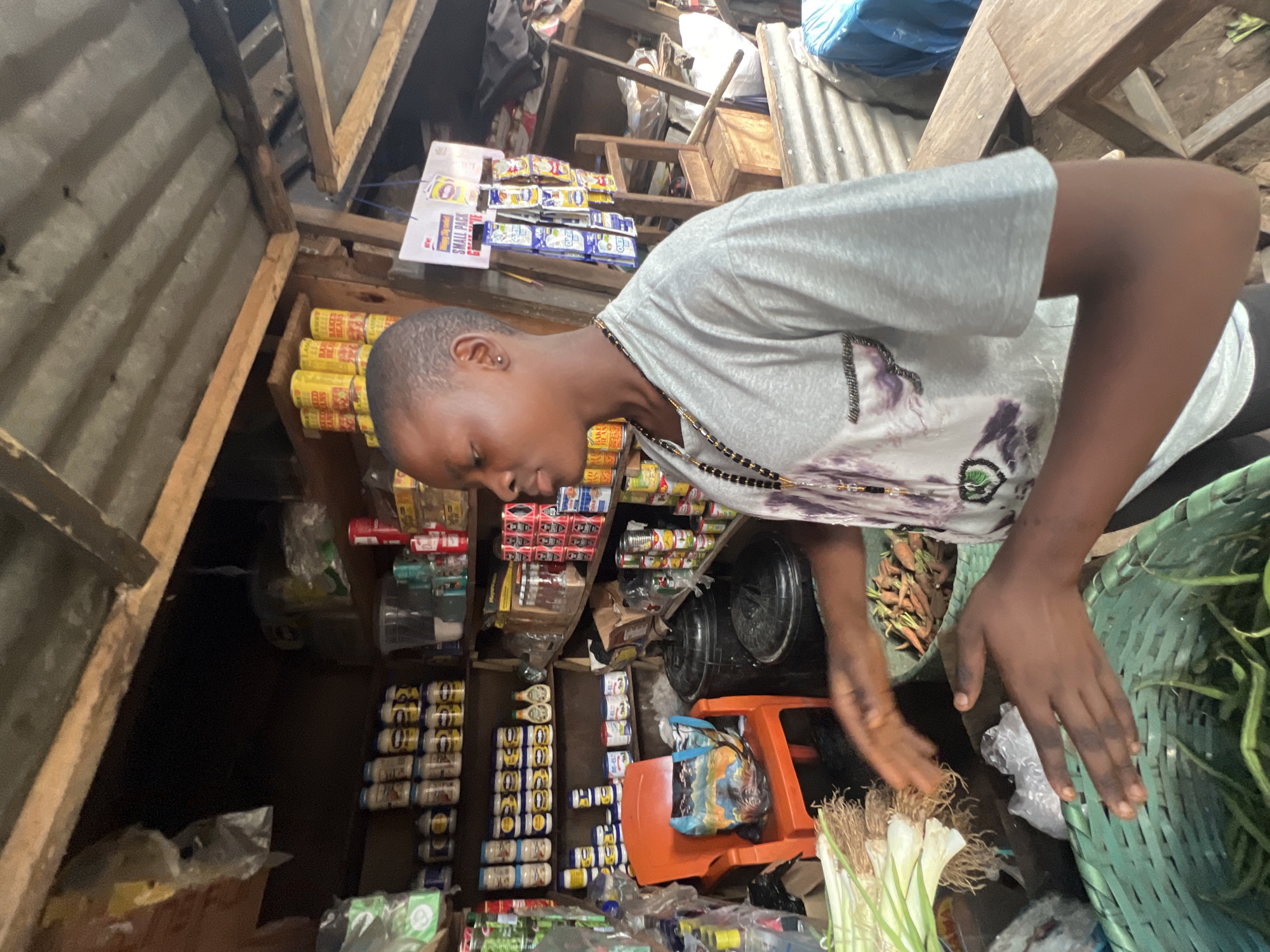 This media file shows a young person with short hair, wearing a grey t-shirt with a purple design and a beaded necklace. They are in what appears to be a small market stall or shop. The stall is filled with various products, including cans stacked on shelves, bags of goods, and some fresh produce like carrots and leeks in baskets. The person is looking down and reaching out with their hands, possibly examining or selecting items.
The setting is rustic, with wooden structures and corrugated metal visible. The lighting suggests it could be daytime. The overall impression is that of a bustling, local marketplace. There is some visible text on product packaging, but it is mostly unreadable due to the resolution and angle. The scene captures a moment of interaction with the goods in the stall.
