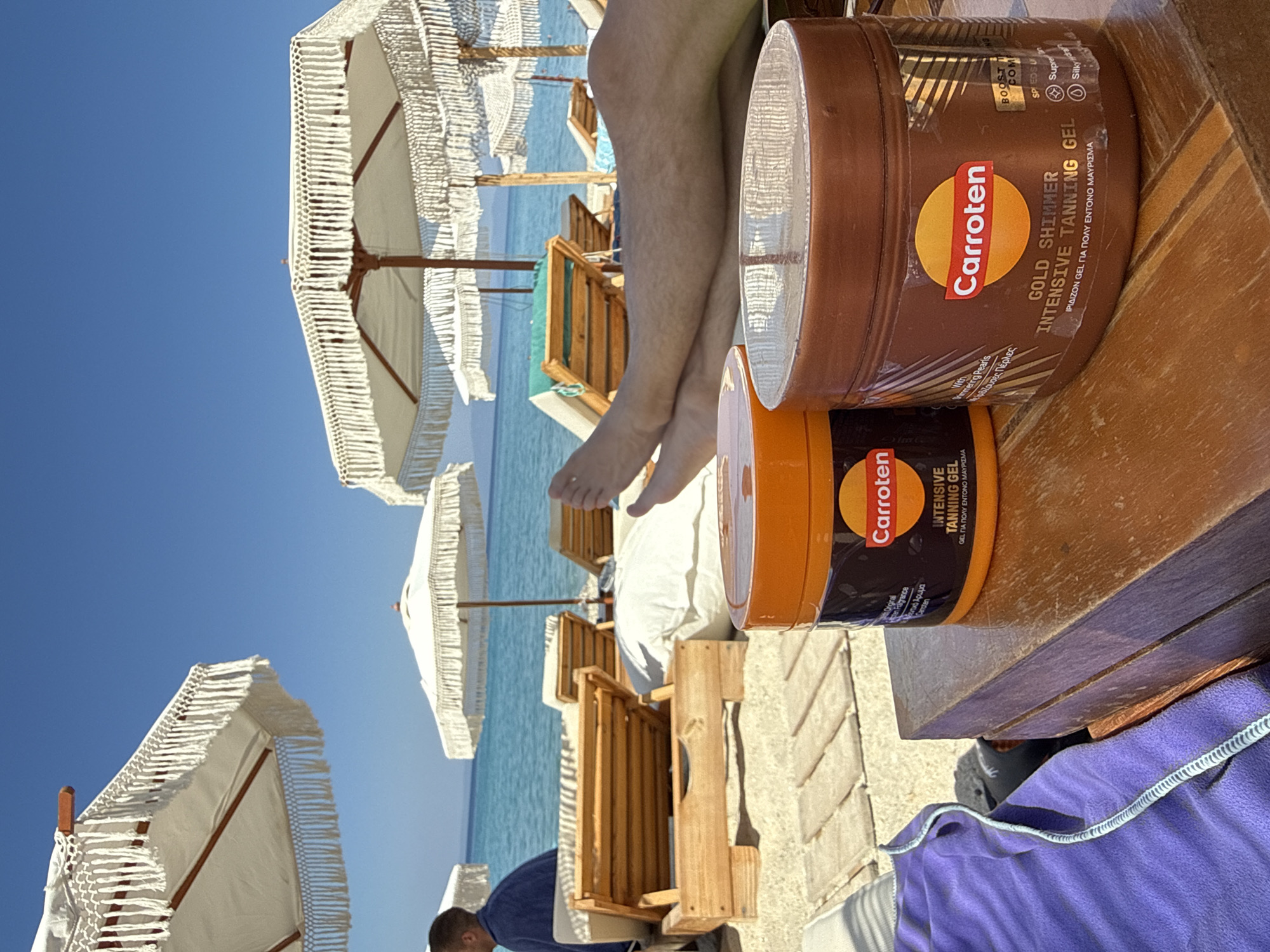 In a sun-drenched scene at a beach in Pallini, Greece, a person is relaxing on a wooden lounger, partially covered by a purple towel. The focus is on two jars of Carroten tanning gel, one labeled "GOLD SHIMMER INTENSIVE TANNING GEL" and the other "INTENSIVE TANNING GEL." The jars are positioned on a wooden surface, suggesting they are readily accessible for sunbathing.
Behind the loungers and jars, pristine white beach umbrellas with fringed edges dot the landscape. The turquoise waters of the Aegean Sea are visible in the background, indicating a clear, sunny day. The presence of the umbrellas and the sea suggests a typical day at the beach, likely during the summer months. The overall atmosphere is one of relaxation and enjoyment of the sunny Greek weather. The visible text on the tanning gel jars clearly indicates the product's purpose for tanning and its brand. The scene evokes a sense of vacation and leisure.