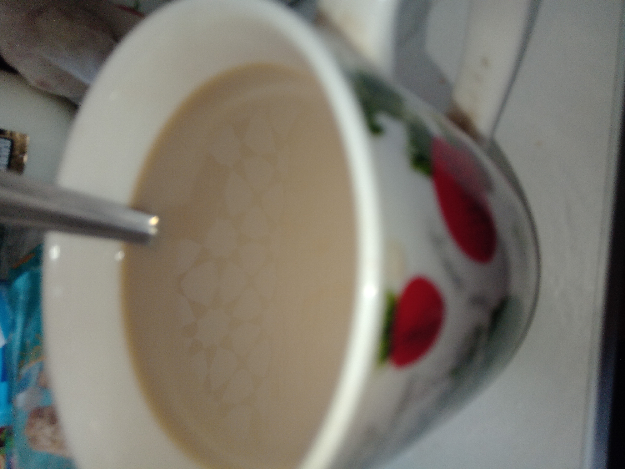 A close-up, slightly blurry photograph shows a white mug filled with a light brown beverage, likely coffee or tea, with a metal spoon resting in it. The mug has a decorative pattern of red strawberries and green leaves on its exterior. The background is out of focus, but appears to be an indoor setting with some items on a table or shelf. There is no information to suggest this image is located in Buguruslan, Russia.