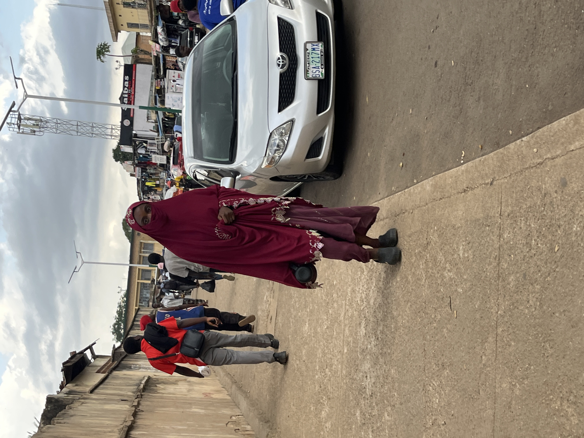 The image depicts a street scene in Jos, Nigeria, under an overcast sky. A woman wearing a dark red hijab and a matching dress that covers her face, exposing only her eyes, walks towards the camera. She carries a dark object in her right hand. To her left, a silver Toyota Corolla is parked on the road, its license plate reading "BSA 217MX". In the background, several people are walking on the sidewalk, some carrying bags. Buildings line the street, with shops and a prominent cellular tower visible. A man in a red shirt and grey pants is walking away from the camera, with a black bag slung over his shoulder. The environment appears to be a busy urban area with a mix of pedestrians and vehicles.