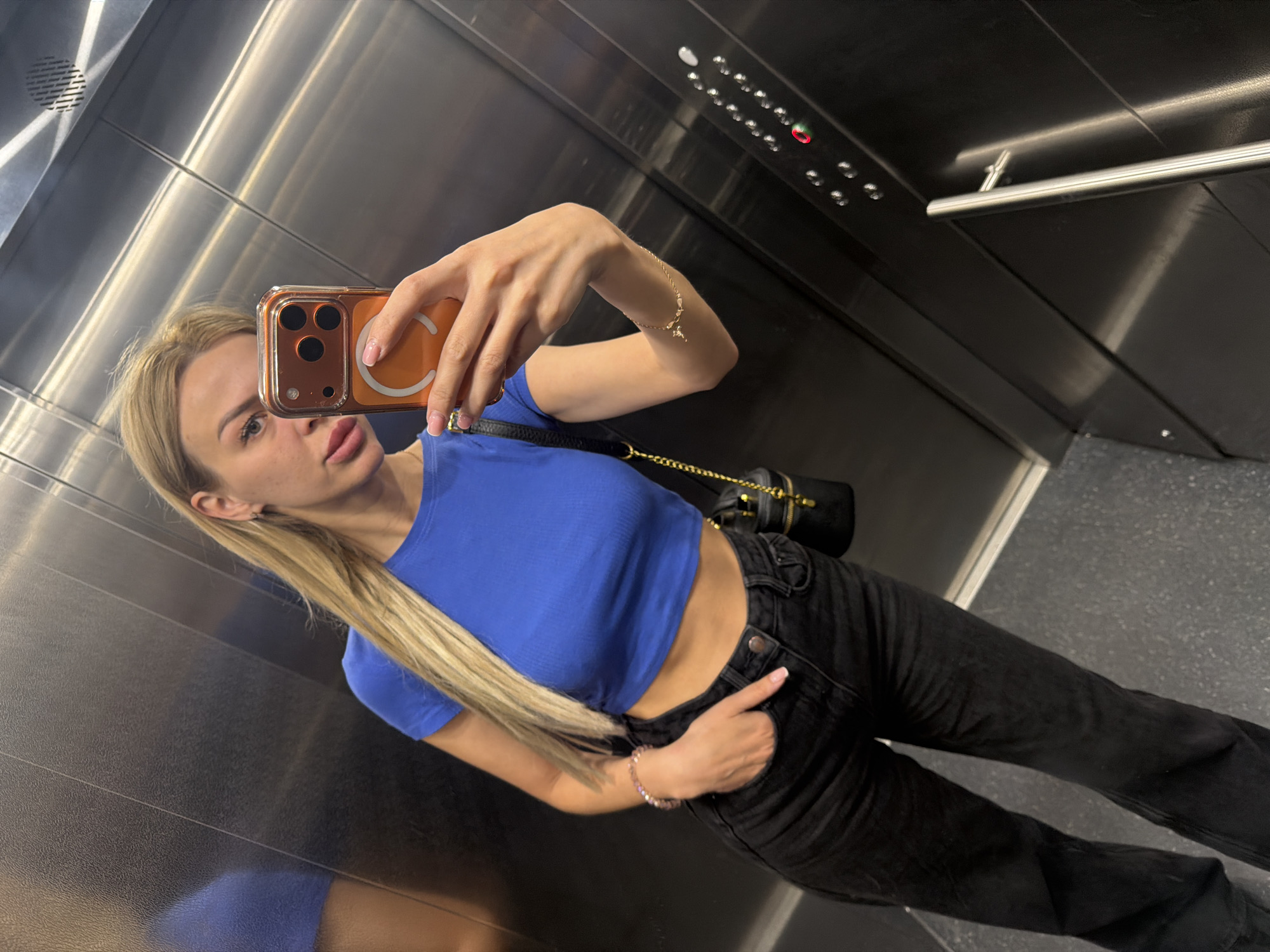A woman takes a mirror selfie in an elevator. She has blonde hair and is wearing a blue crop top and black jeans. She is holding an orange phone with a camera and has a black bag slung over her shoulder. The elevator has silver walls and a button panel with a red light on. The scene is likely within the city of Kyiv, Ukraine, given previous image context.