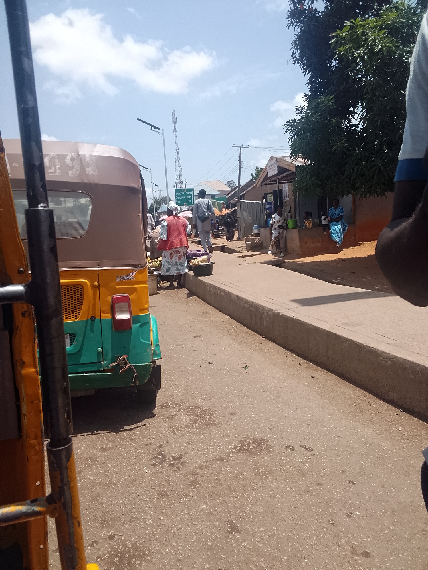The image depicts a street scene in Bukuru, Nigeria. A three-wheeled vehicle (likely a "Keke") is prominent in the foreground. Further ahead, people are seen walking along the sidewalk and engaging in commerce. Buildings line the street, and signage indicates distances to nearby towns like Manchok, Ganawuri, and Vom. The weather appears sunny.