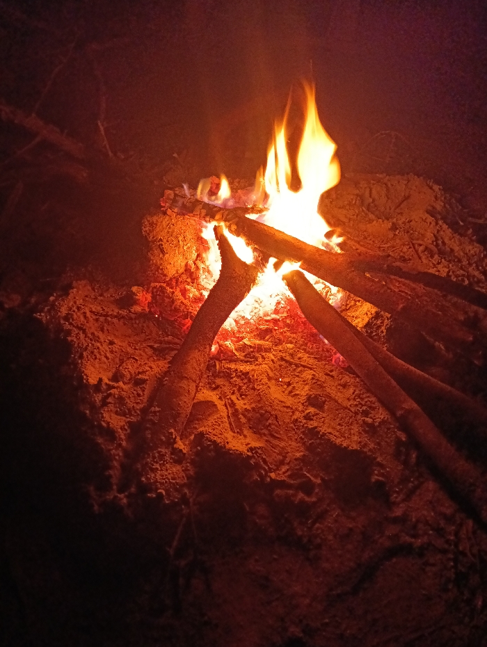 A bonfire is centrally depicted on sandy or dirt ground at night in Kyamatu, Kenya. The fire consists of multiple dark wooden logs or branches arranged over a core of intensely glowing orange and red embers. Vibrant yellow and orange flames rise vertically from the burning wood. The immediate surrounding ground is illuminated by the fire's light, revealing reddish-brown soil or sand with visible textures. Areas further from the flame transition into deep shadow. No human figures or additional objects are discernible within the frame beyond the fire and its immediate environment.