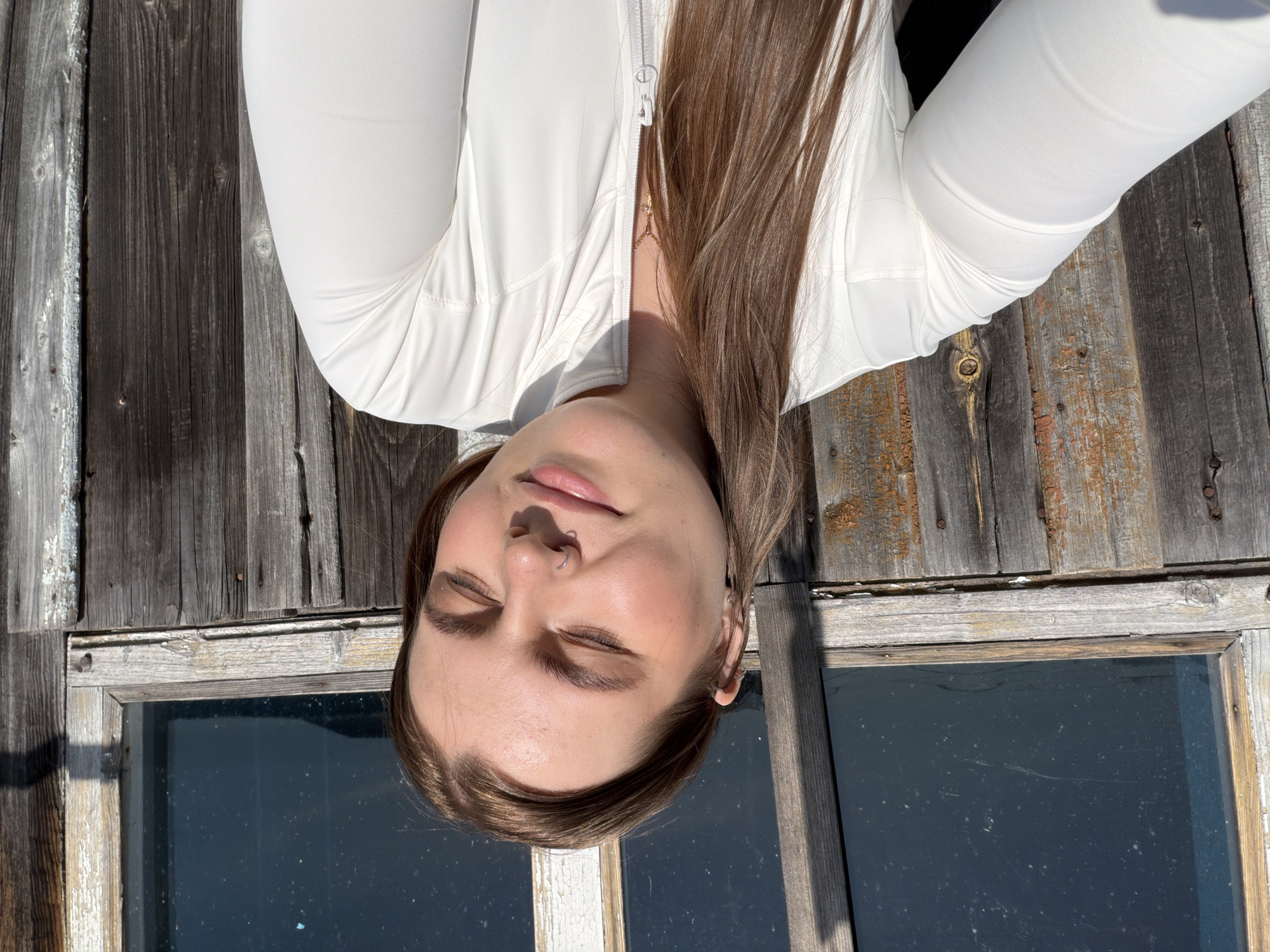 A young woman with light brown hair and fair skin is lying on a weathered wooden surface, potentially a dilapidated structure with a window pane visible below her head. She is wearing a white, long-sleeved top or jacket that is partially zipped up. Her eyes are closed, and she has a nose ring on her left nostril. Her location and whether she is in Чумна, Russia, is not discernible from the visual data of the image.