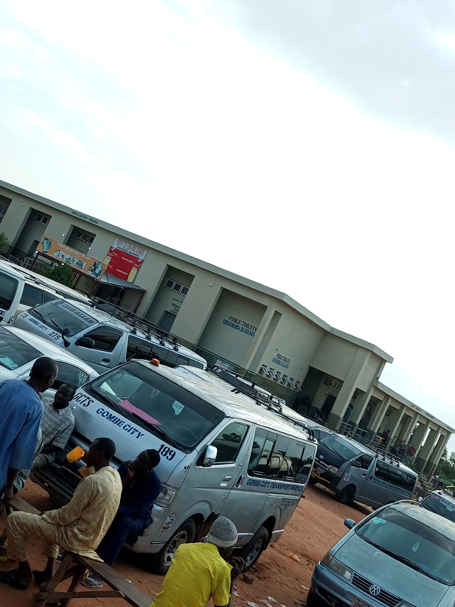 In Gombe, Nigeria, a cluster of vehicles, primarily minibuses and cars, are parked on a dirt lot adjacent to a beige, two-story building with a covered walkway and numerous windows. Above the walkway, text reads "ABDULLAHI MAI HINARAS". Several signs are affixed to the building's facade, including one advertising "YUSUF DELOW BEST SELLER" and another with red accents that appears to be a commercial advertisement. People are present among the vehicles; one man in a yellow top is seated on a wooden bench, holding a yellow cup, while others stand or sit near the minibuses. The minibuses are silver and have various markings, including "GOMBE CITY 275" and "189" on one of them. Another minibus bears the text "GOMBE CITY TRANSPORT SERVICE". A light blue sedan is parked in the foreground. The sky is overcast with a pale, light blue hue. The ground is rough, reddish-brown dirt.